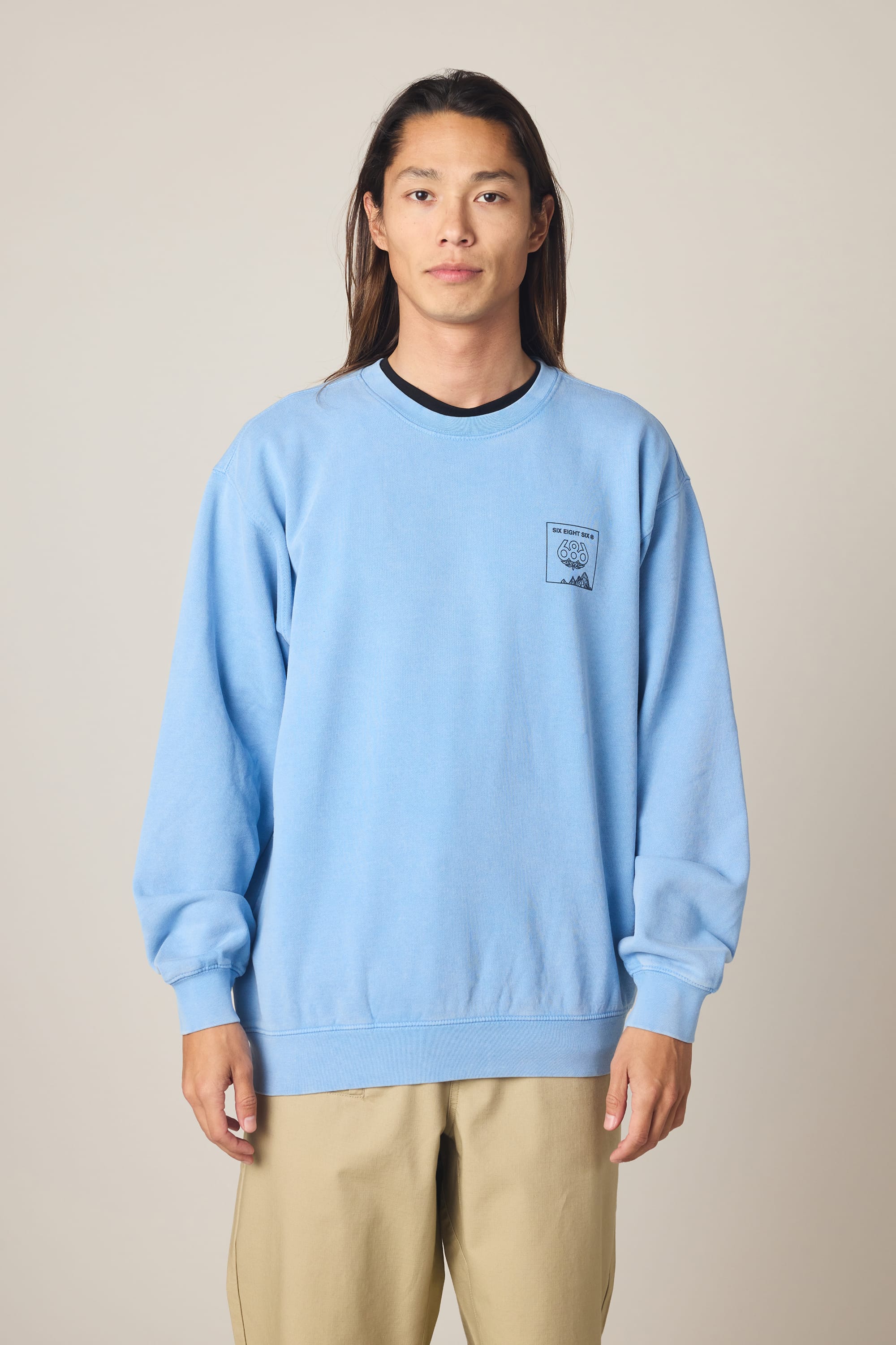 A person with long brown hair stands facing forward, wearing the 686 Men's Mixed Relaxed LS Crewneck by 686 in light blue and beige pants, against a plain light background.