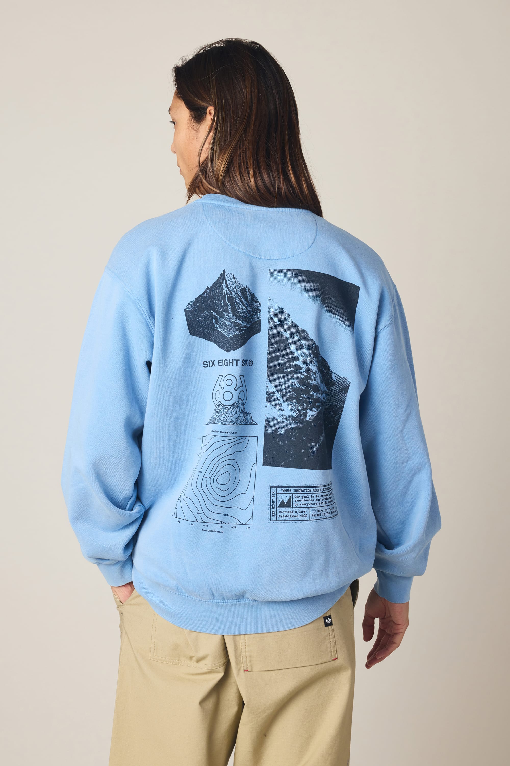 A person wearing the 686 Men's Mixed Relaxed LS Crewneck—a light blue 100% cotton sweatshirt by 686 featuring black-and-white mountain imagery, topographic map graphics, and text on the back—stands against a plain background.