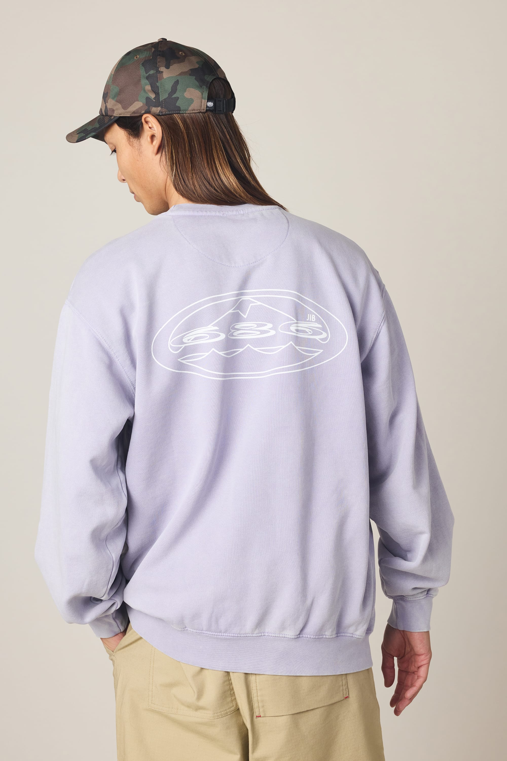 A person wearing a camo cap, beige pants, and the 686 Men's Mixed Relaxed LS Crewneck by 686—100% cotton, light purple with a white abstract back design—stands facing away from the camera against a plain background.