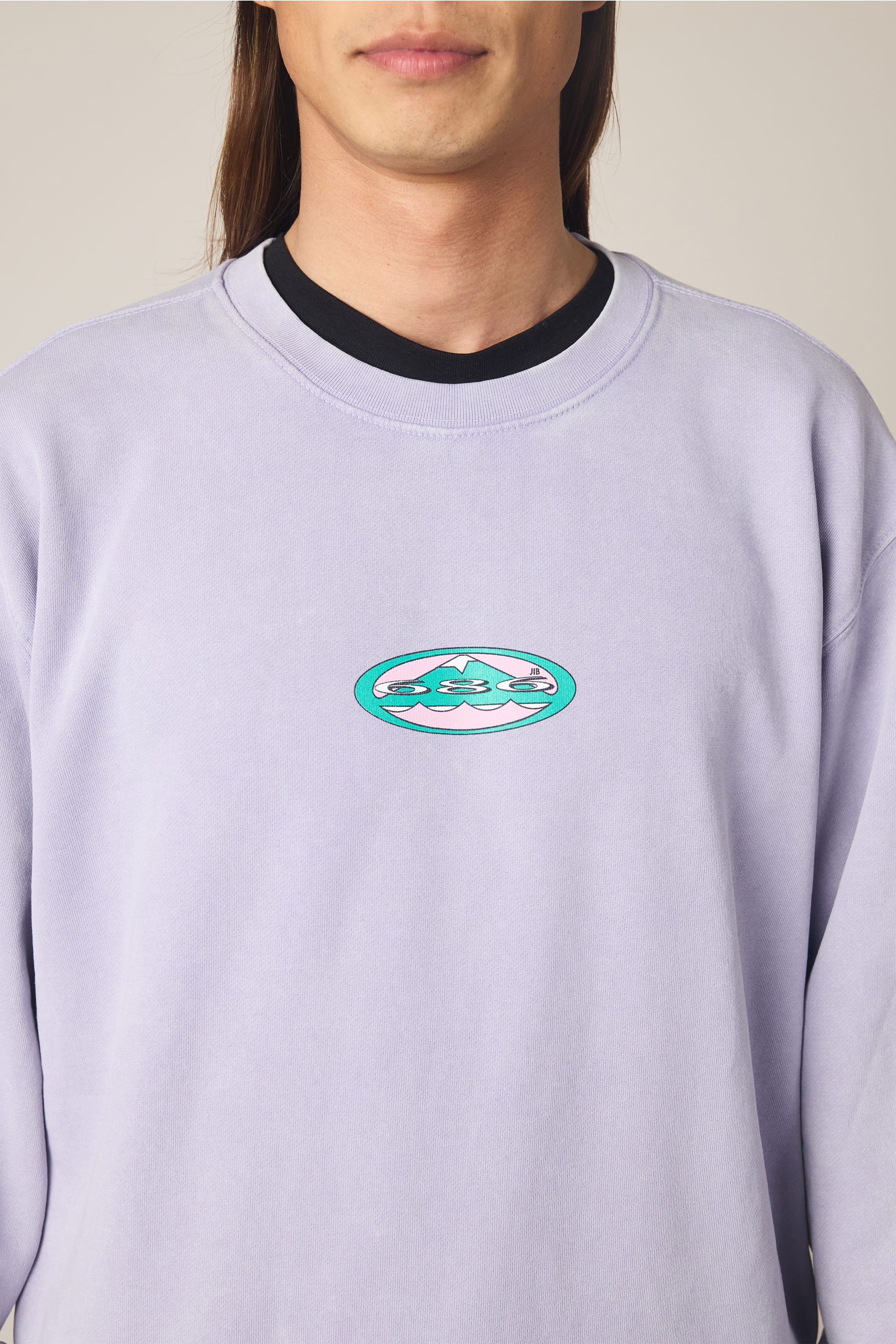 A person wearing the 686 Men's Mixed Relaxed LS Crewneck, a light purple long-sleeve t-shirt from 686, featuring a small oval-shaped logo with turquoise and white abstract shapes on the center front. Only the upper torso and lower face are visible, emphasizing the relaxed fit and 100% cotton comfort of this men's shirt.