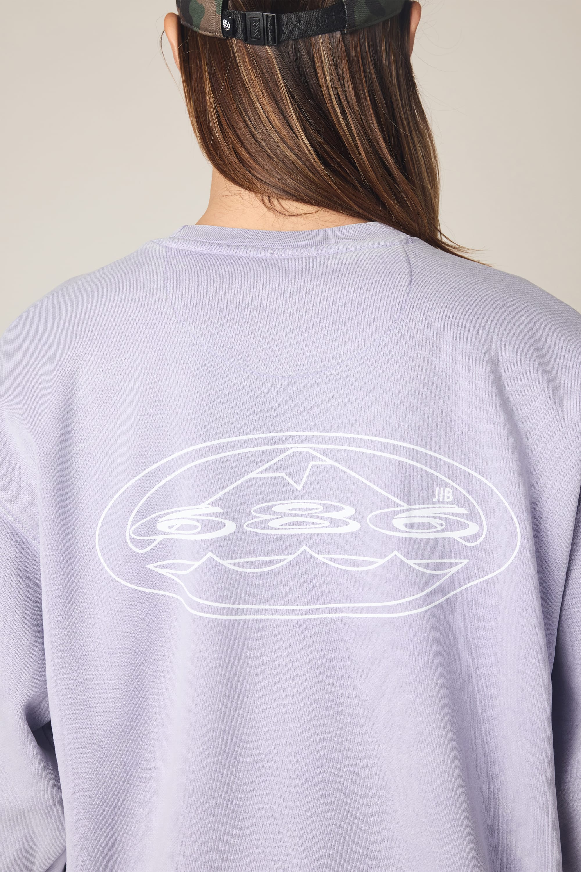 Sure! Here’s the revised sentence using your product data: A person with long hair is shown from behind wearing the 686 Men's Mixed Relaxed LS Crewneck—a light purple, 100% cotton sweatshirt featuring a white abstract graphic and the initials JIB on the back—paired with a dark camouflage cap.
