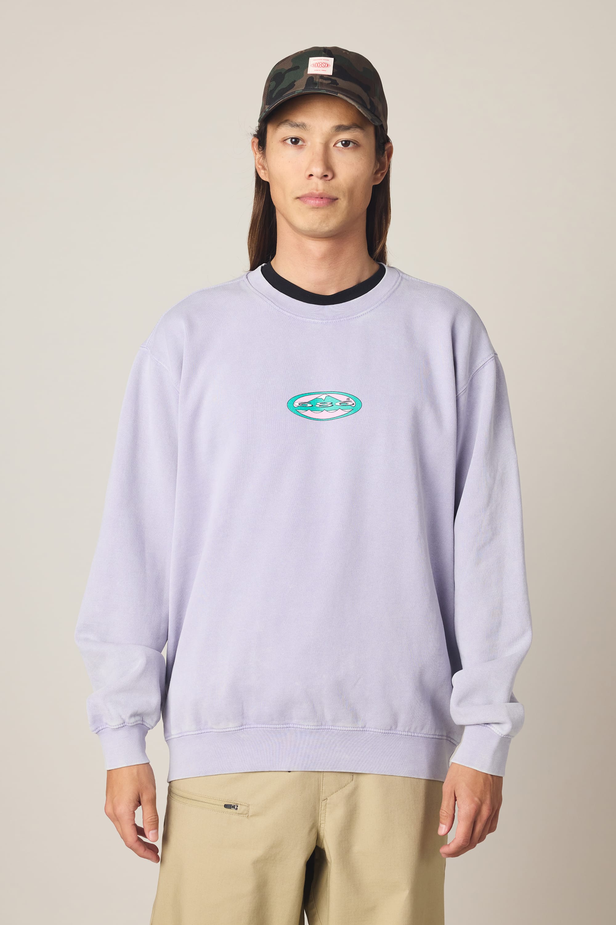 A person stands against a plain light background wearing the 686 Men's Mixed Relaxed LS Crewneck in light purple, featuring a small blue and green oval logo at the center, paired with tan pants and a camouflage baseball cap.
