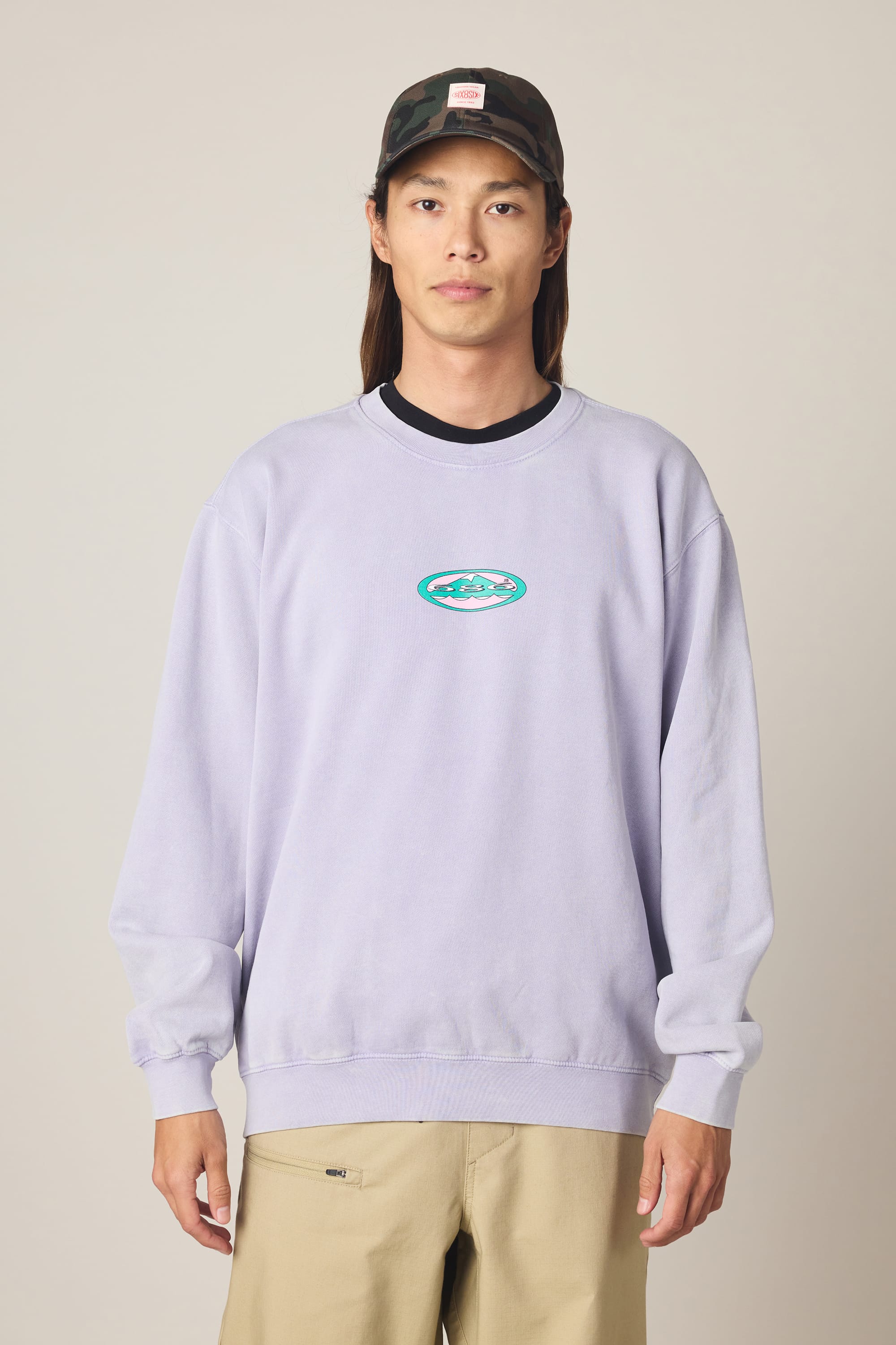 A person wearing the 686 Men's Mixed Relaxed LS Crewneck, a light purple 100% cotton sweatshirt featuring a small oval logo, along with khaki pants and a camouflage cap, stands against a plain light background looking at the camera with a neutral expression.