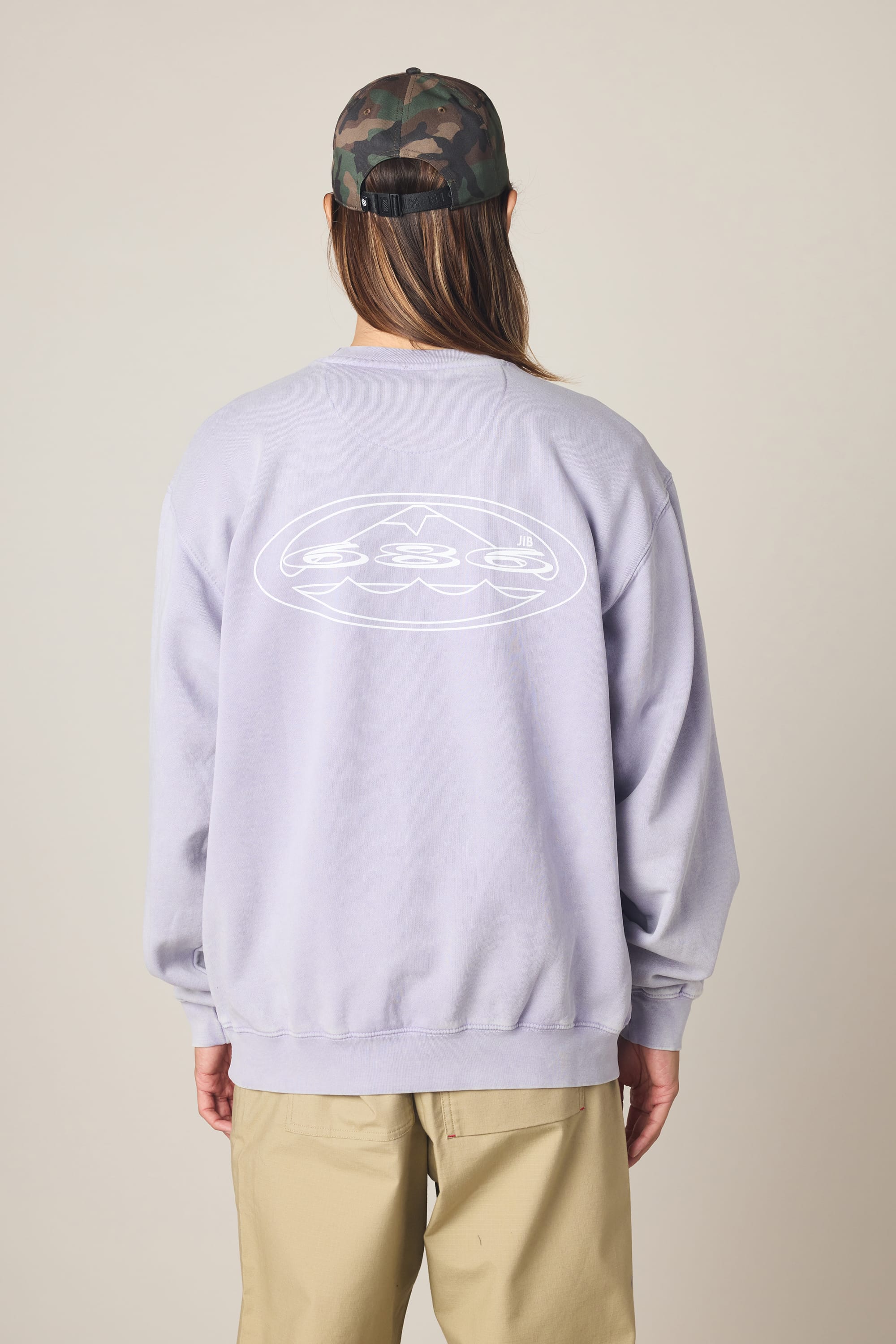 A person with long hair is seen from behind against a plain background, wearing a camo cap, beige pants, and the 686 Men's Mixed Relaxed LS Crewneck by 686—a light purple cotton sweatshirt featuring a white abstract design on the back.