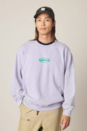 A person with long brown hair wears a black cap, beige pants, and the 686 Men's Mixed Relaxed LS Crewneck by 686—a light purple 100% cotton sweatshirt with a small embroidered logo—standing against a plain light background.