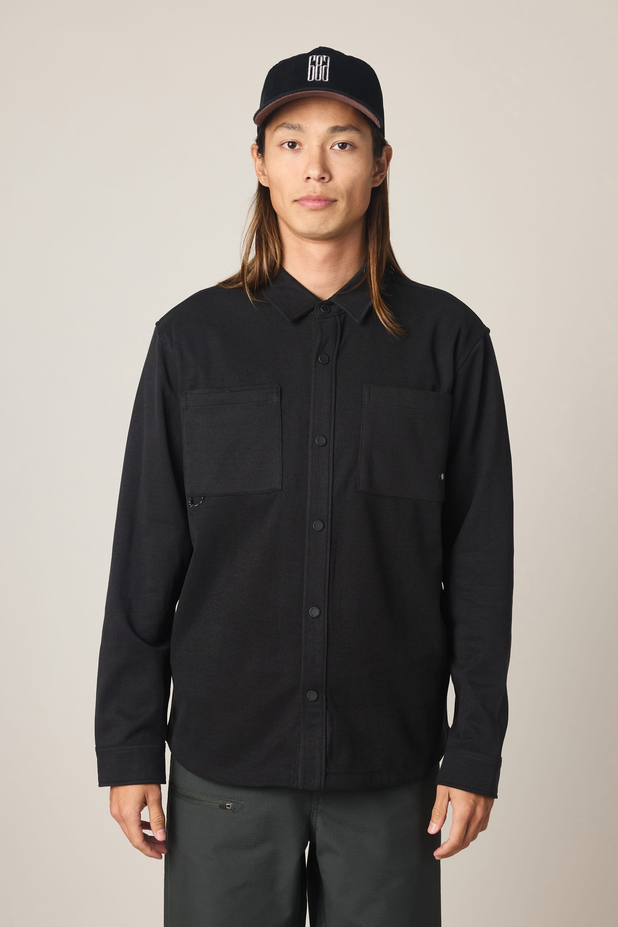 Wearing the 686 Men's Whisper Knit Flannel—black, button-up, with two chest pockets—a person with long hair pairs it with dark pants and a black baseball cap featuring a white logo, standing against a plain light background.