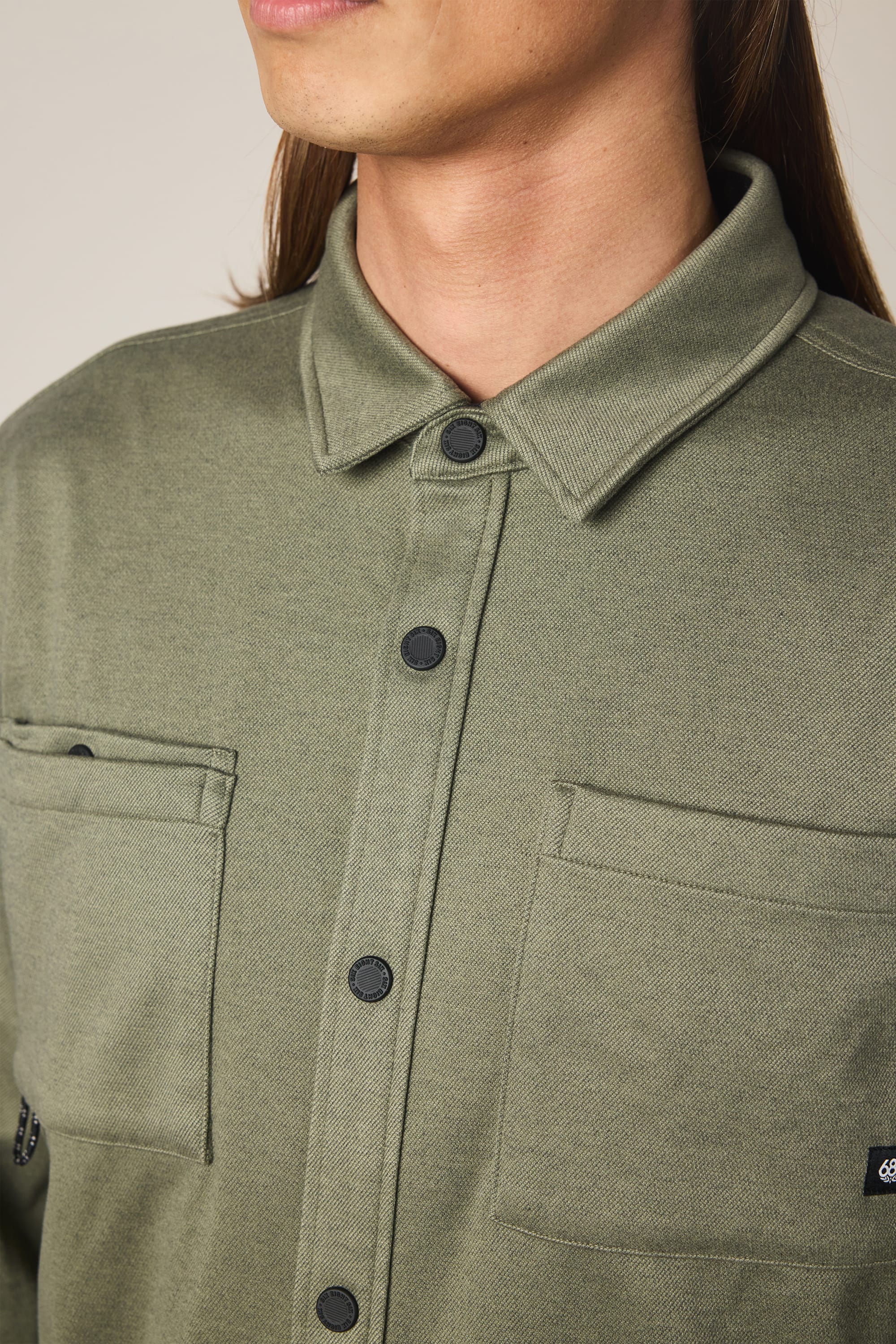 Close-up of a person with long brown hair wearing the 686 Men's Whisper Knit Flannel in olive green, featuring two chest pockets and black buttons, facing slightly to the left. Brand: 686.