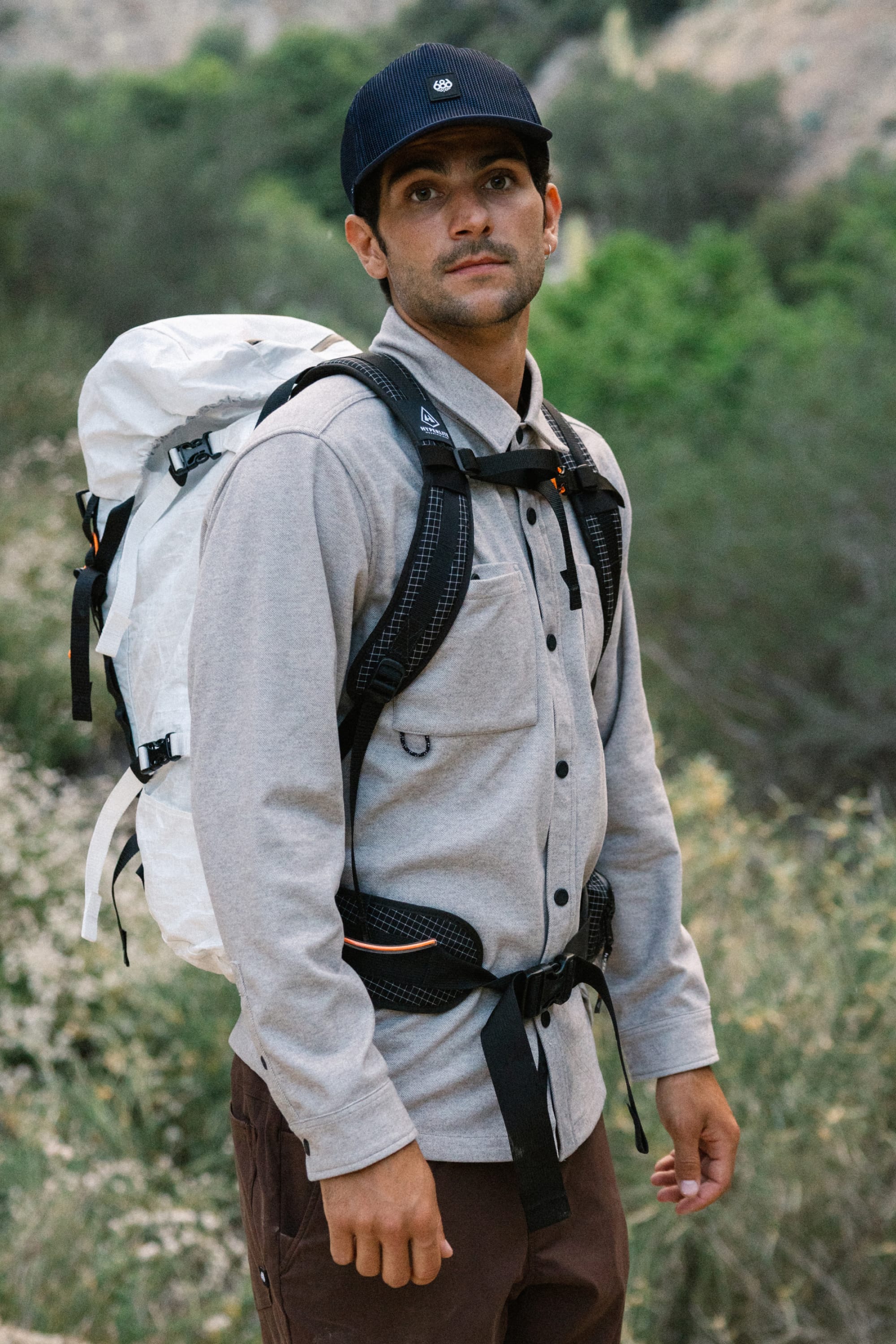 A man wearing the 686 Men's Whisper Knit™ Flannel by 686, along with a black cap and a white hiking backpack, stands outdoors with greenery and hills in the background.