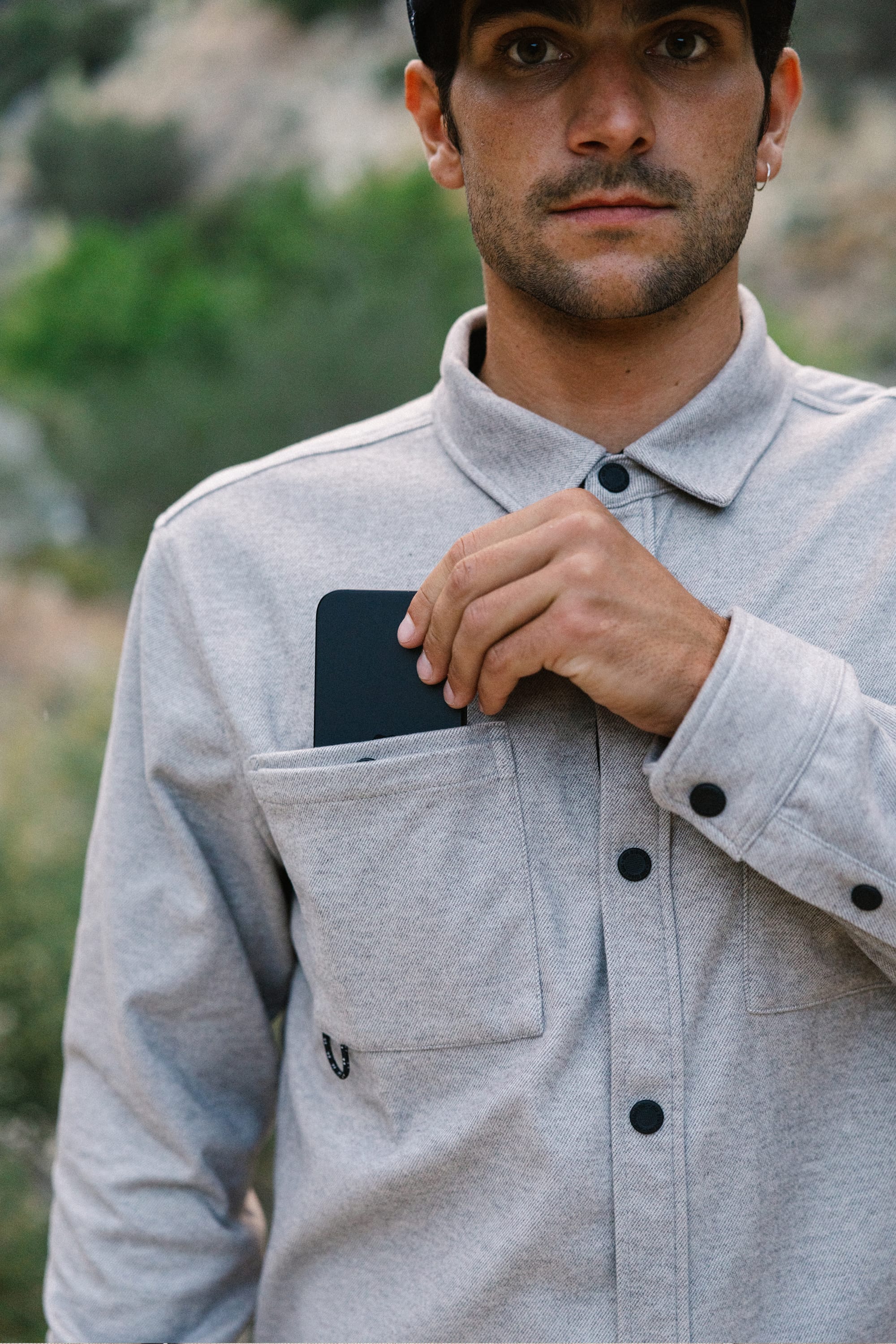 A man wearing the 686 Men's Whisper Knit™ Flannel in light gray puts a black smartphone into his chest pocket outdoors, with blurred greenery in the background.