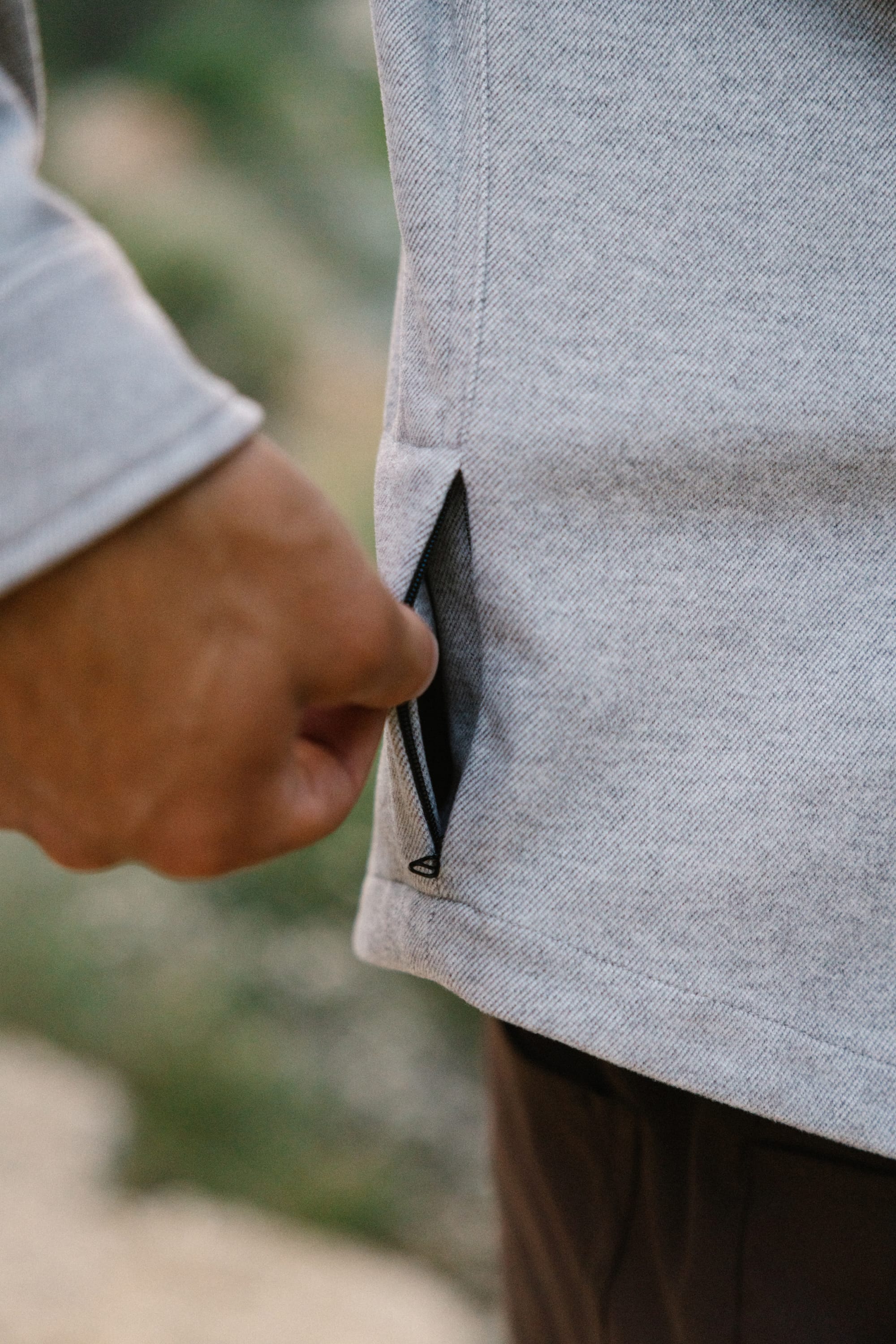A person’s hand pulling on the zipper of a pocket on a light gray 686 Men's Whisper Knit™ Flannel shirt outdoors.