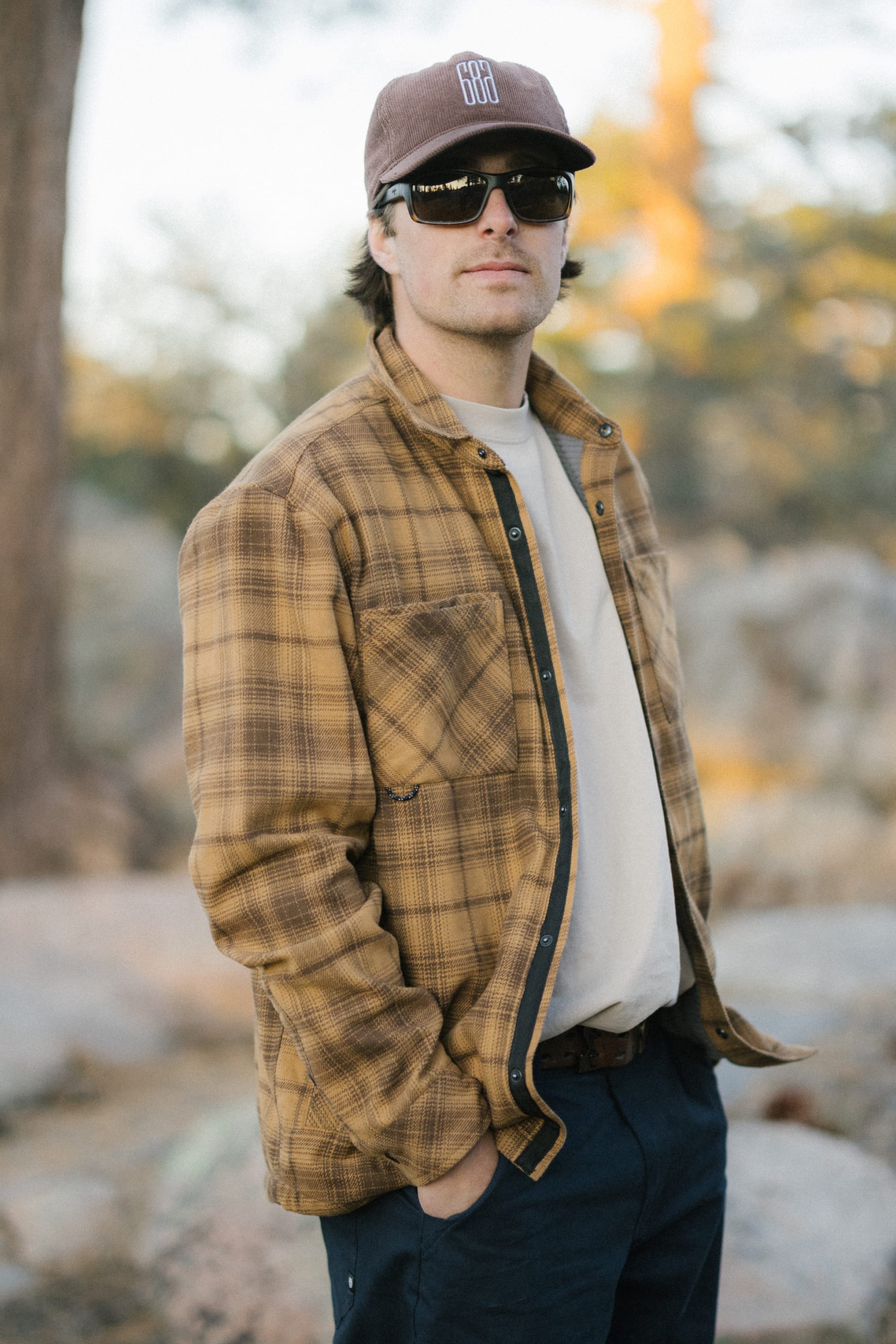 A man wearing sunglasses, a brown plaid jacket, the 686 Men's Heat Grid™ Flannel—a light and comfortable polyester knit shirt by 686—and a brown cap stands outdoors with his hand in his pocket. Trees and rocks are blurred in the background.
