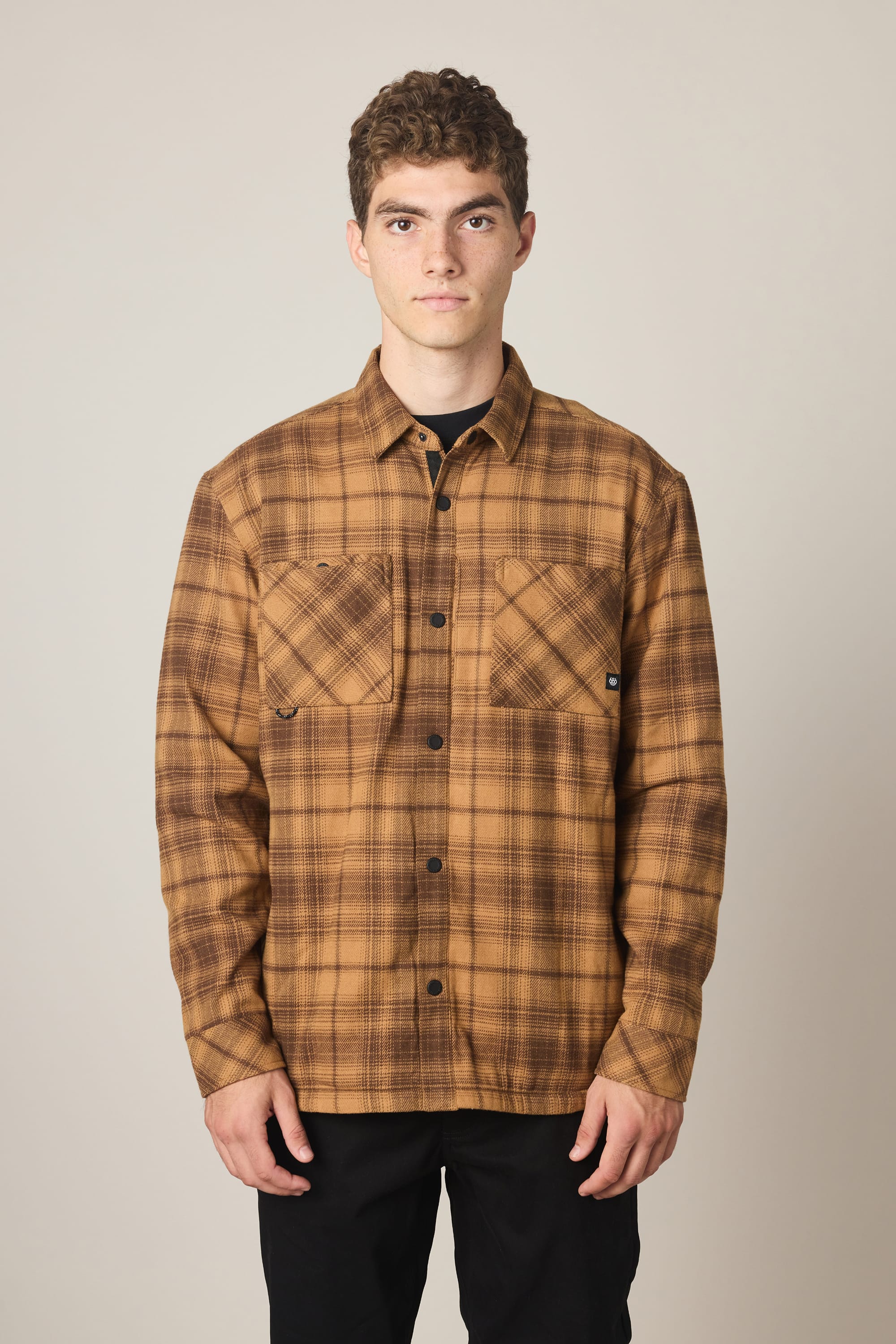 A person stands facing forward against a plain light background, wearing the 686 Men's Heat Grid™ Flannel by 686—a brown plaid shirt made from 100% polyester with two chest pockets—paired with black pants.