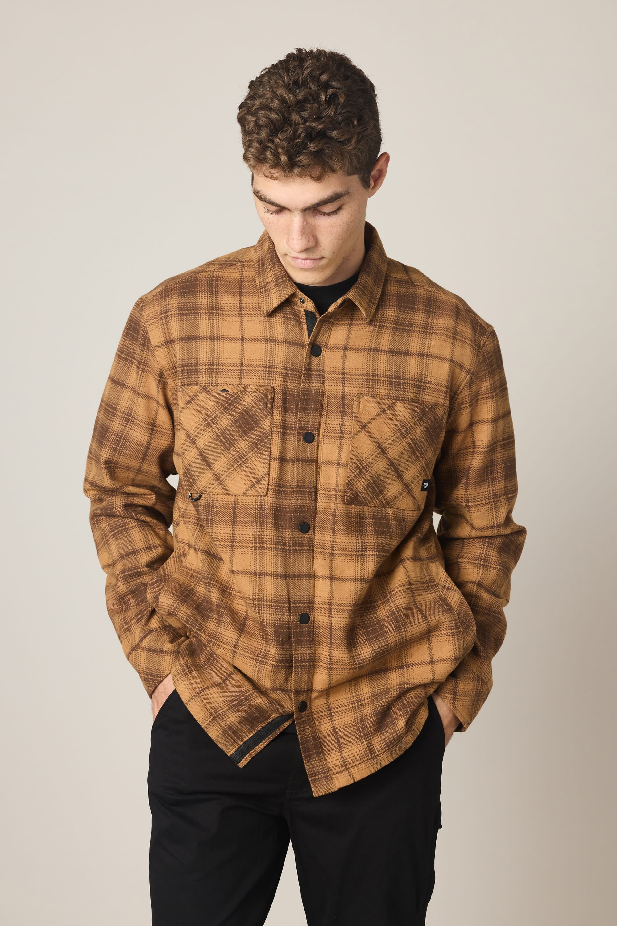A man with short curly hair wears the 686 Men's Heat Grid Flannel by 686, featuring pockets, and stands against a plain light background looking down with his hands in his pants pockets.