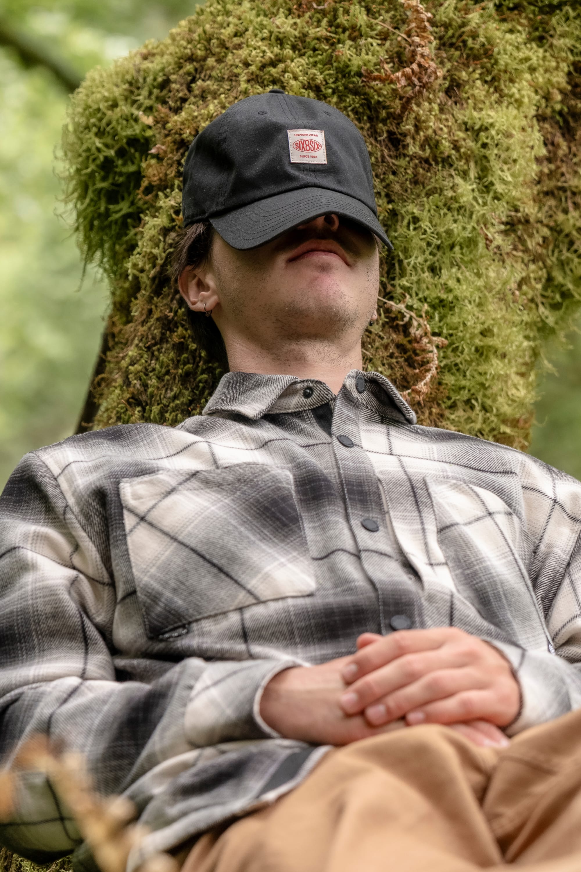 A person wearing a 686 Men's Heat Grid™ Flannel and a black cap is reclining against a mossy tree trunk outdoors, with hands folded and their cap pulled over their eyes, appearing relaxed or asleep.