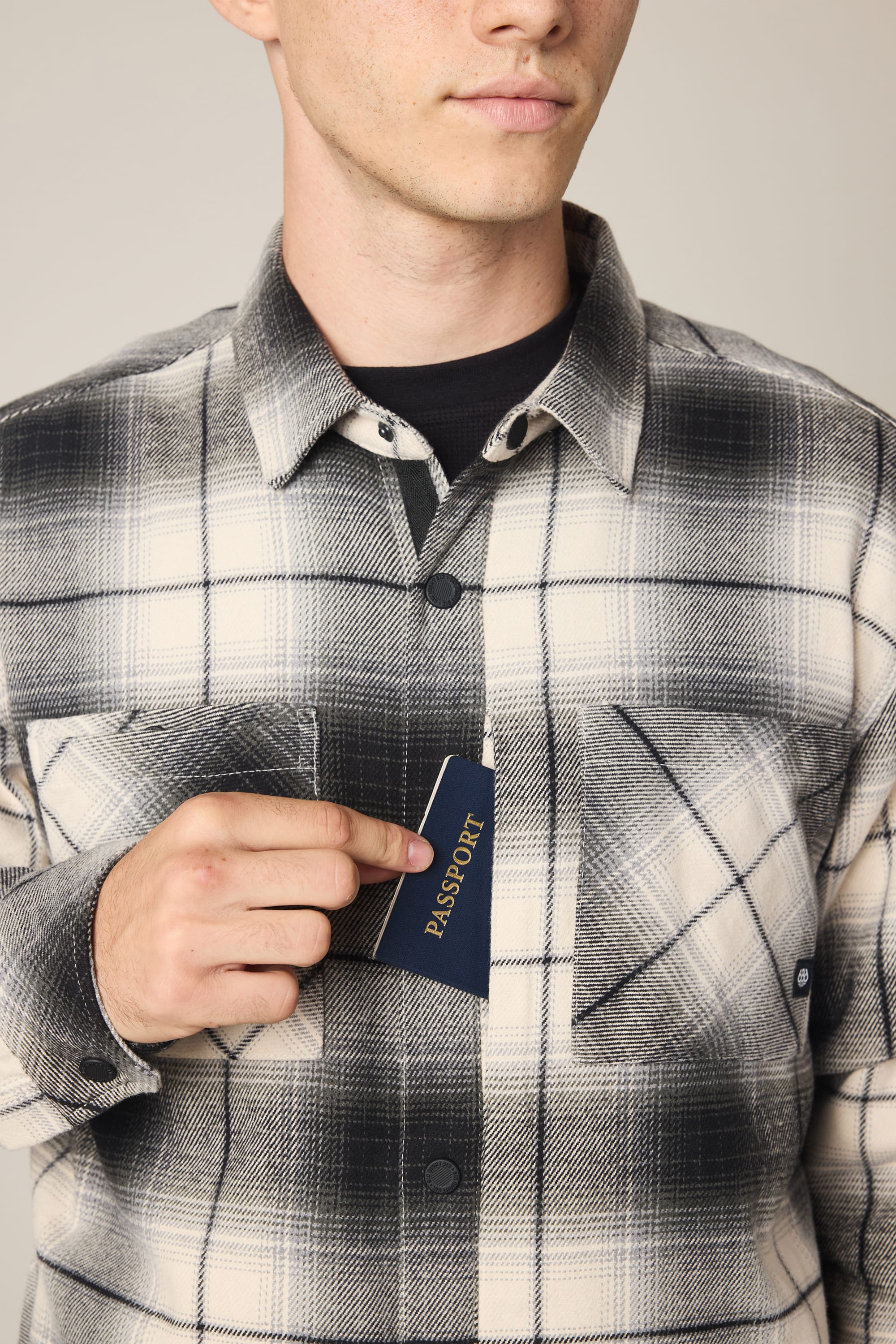 A person wearing a 686 Men's Heat Grid Flannel by 686 with a plaid pattern places a navy blue passport into the shirt’s chest pocket. Their face is partially visible against a plain, neutral background.