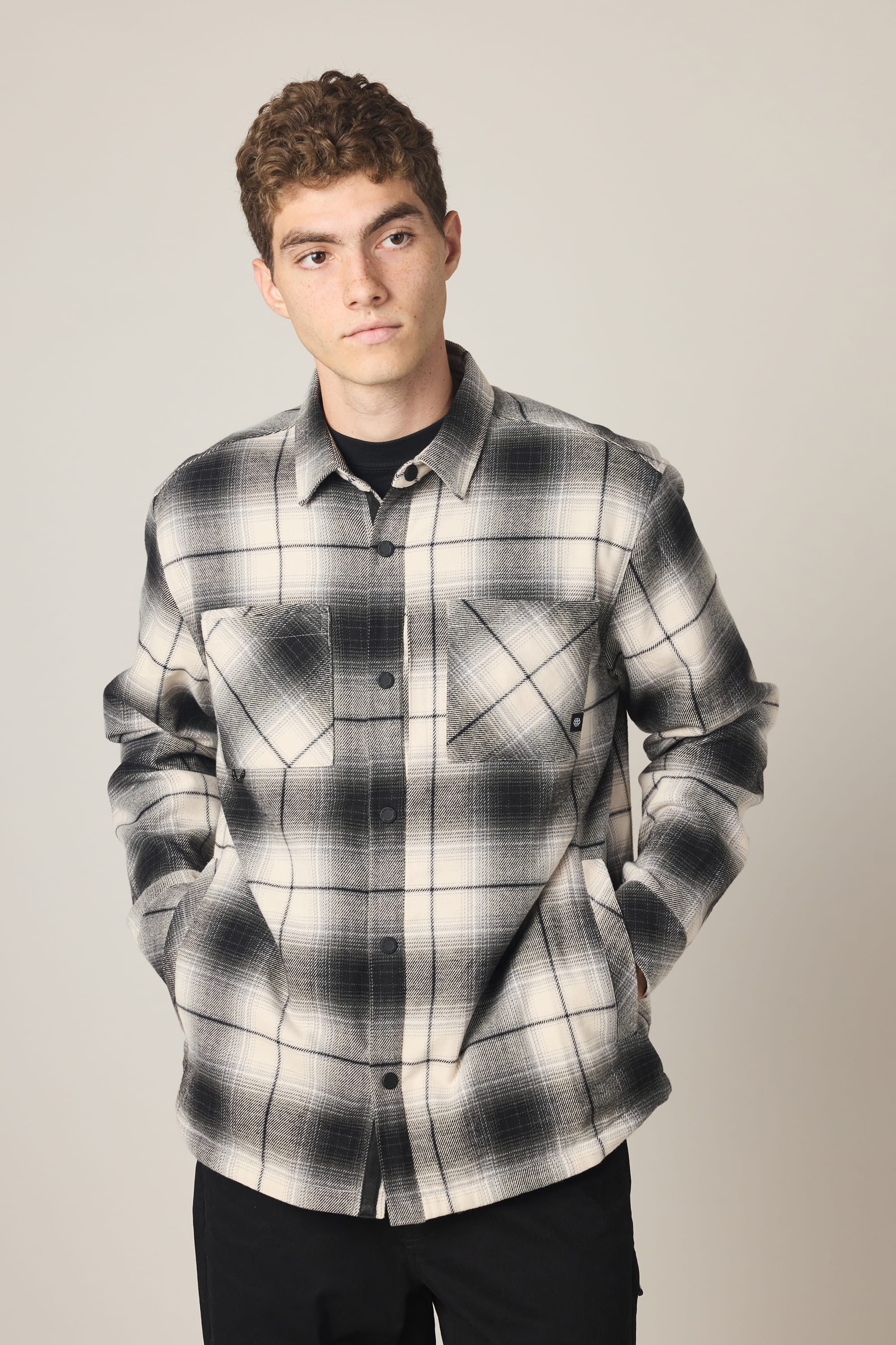 A young man with short curly hair wears the 686 Men's Heat Grid Flannel, a black and white plaid polyester shirt with front pockets. He stands against a plain background, hands in his pockets, looking slightly to the side.