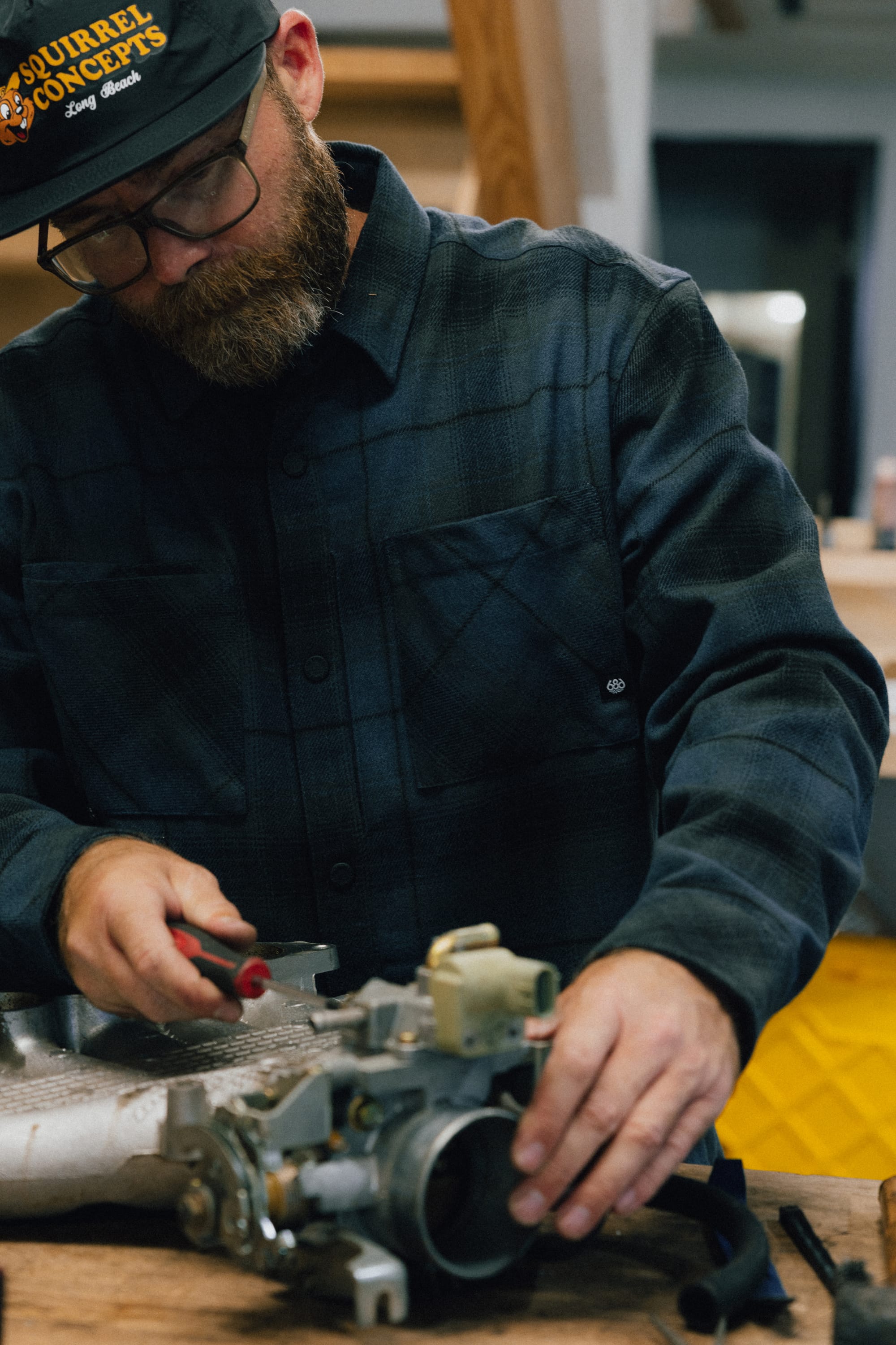 A person wearing glasses and a 686 Men's Heat Grid™ Flannel by 686 uses tools to work on a mechanical part indoors, possibly performing repairs or maintenance.