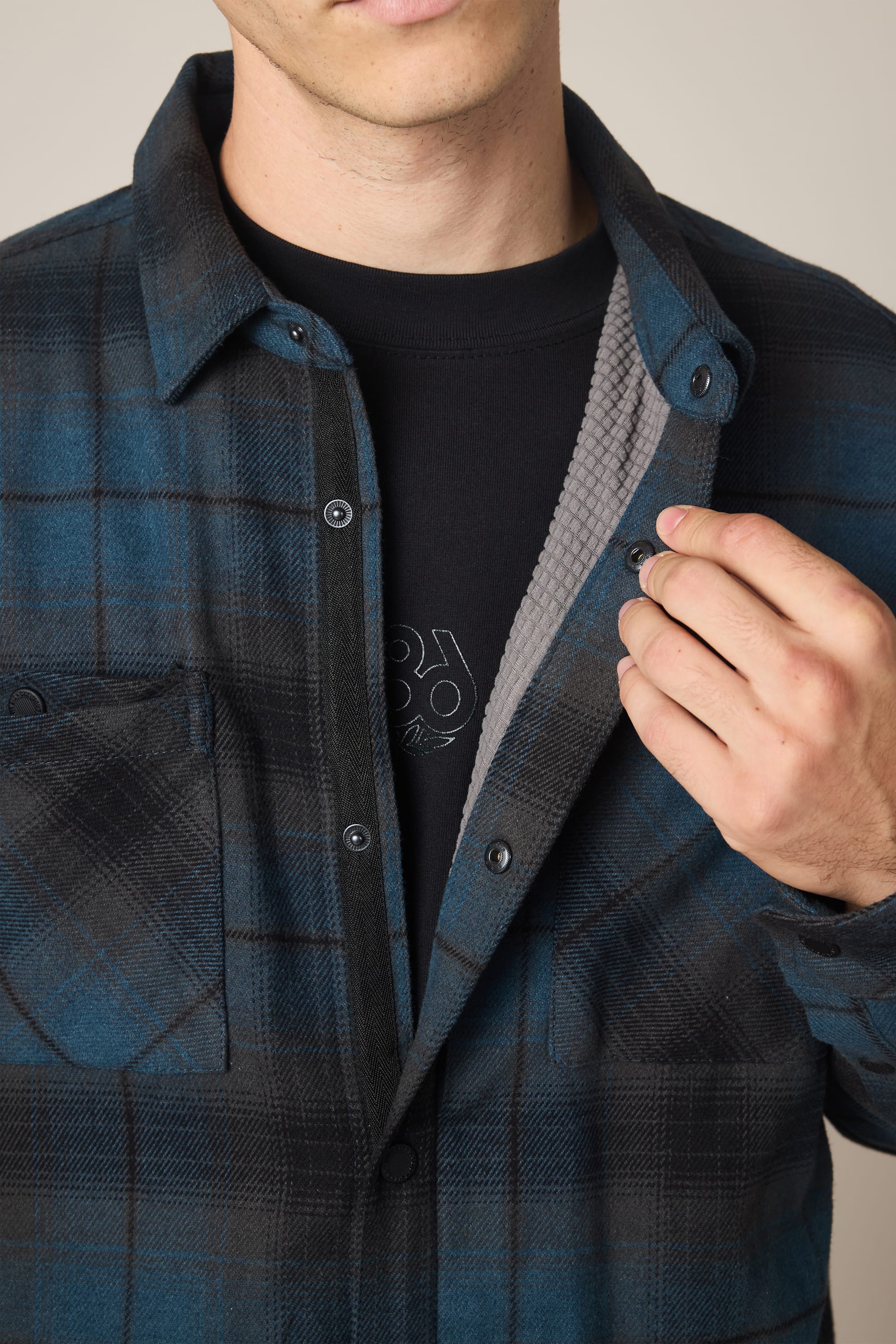 A person wearing the 686 Men's Heat Grid Flannel in blue and black over a black t-shirt holds it open with one hand, displaying its gray inner lining. The face is partially out of frame.