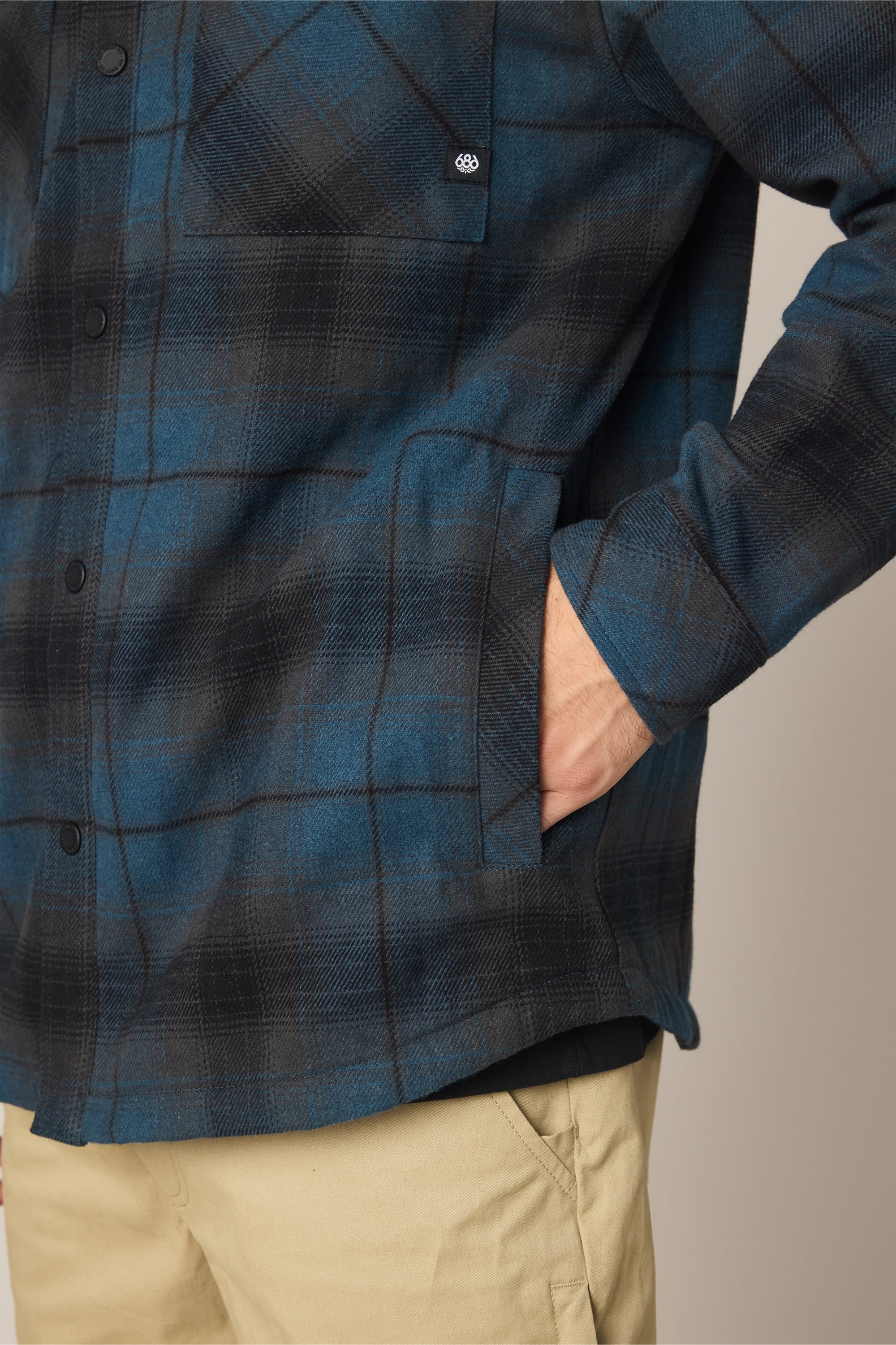 A person wears the 686 Men's Heat Grid Flannel, a blue and black plaid polyester shirt by 686, with their right hand in a side pocket and beige pants. The photo shows the torso, upper legs, and the shirt’s chest pocket.