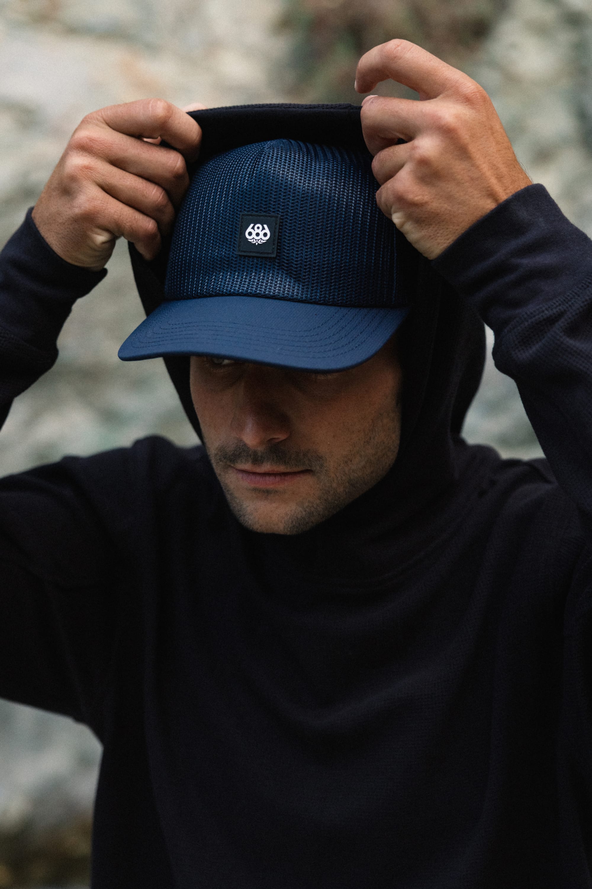 A person wearing a dark hoodie and the 686 High-Pro Knockout Structured 5 Panel Hat by 686, featuring a small white emblem, lifts their hood over their head and looks slightly downward. The background is blurred and outdoors.