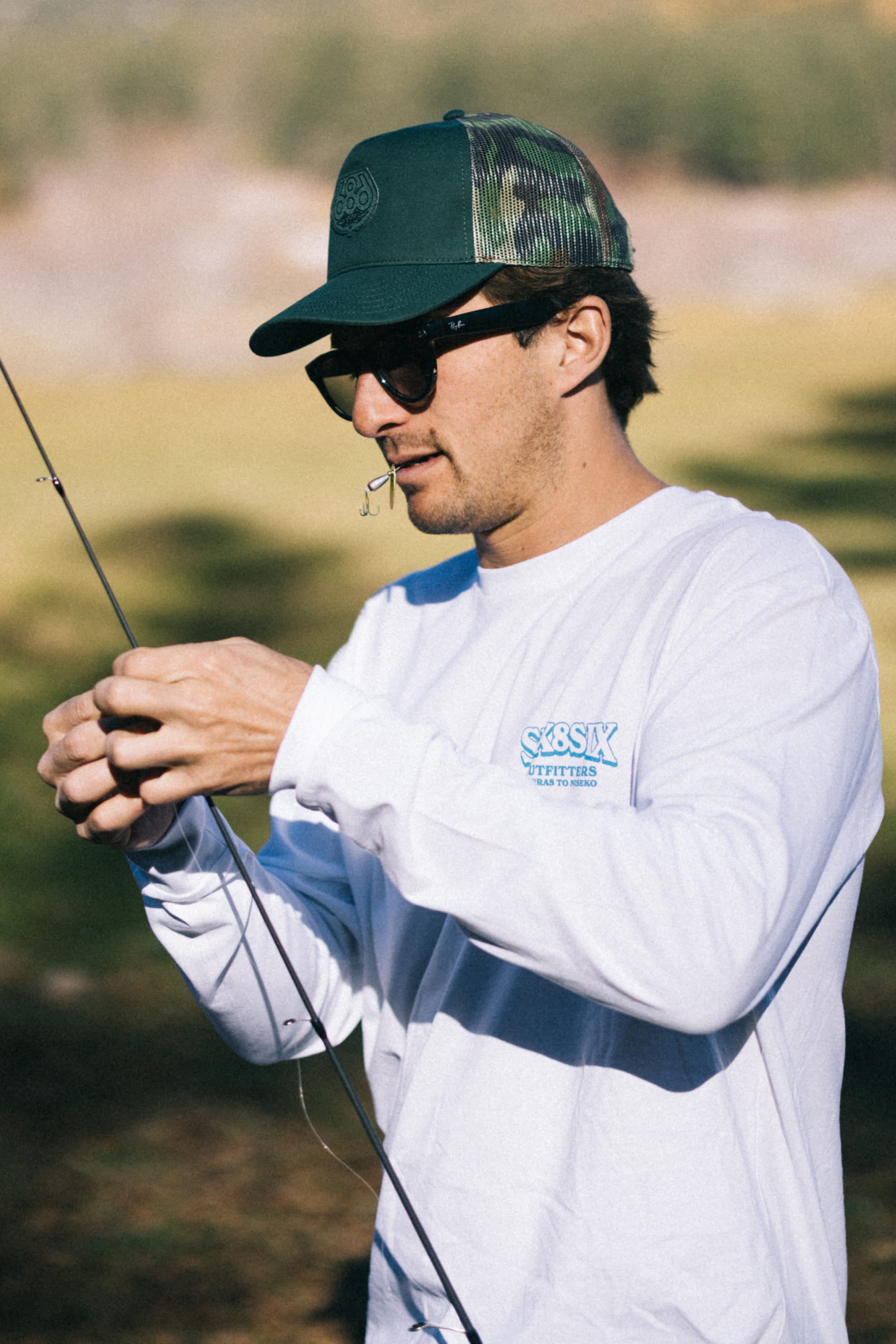A person wearing sunglasses, a 686 Mid-Pro Wreath Unstructured 5 Panel Mesh Hat by 686, and a white long-sleeve shirt works on a fishing line outdoors, holding the line while using their mouth to steady a fishing hook.