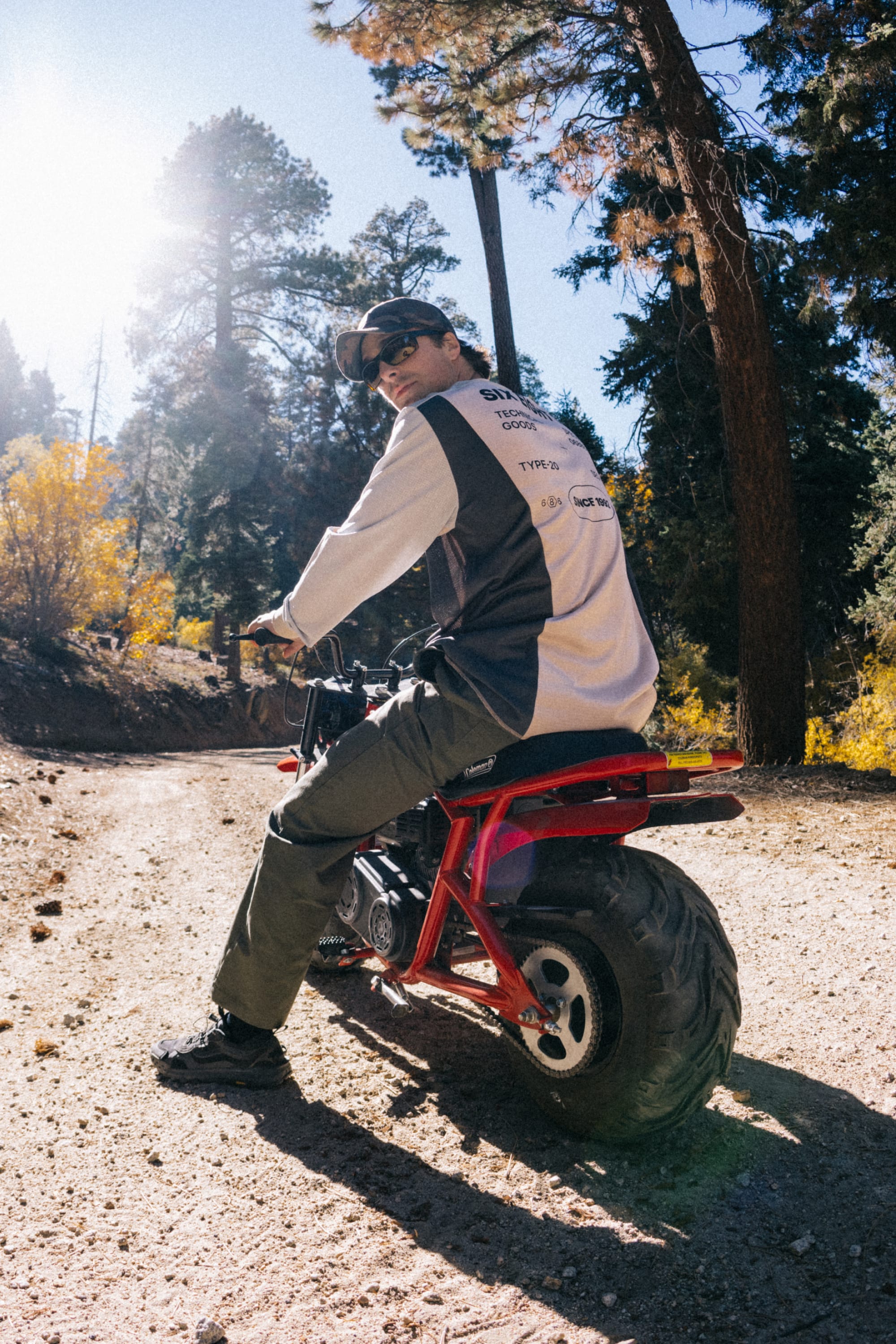 Wearing sunglasses, a 686 Low-Pro Unwork Unstructured 6 Panel Hat by 686, and a long-sleeve shirt, a person sits on a red mini bike along a dirt path in a sunlit forest, glancing back over their shoulder.