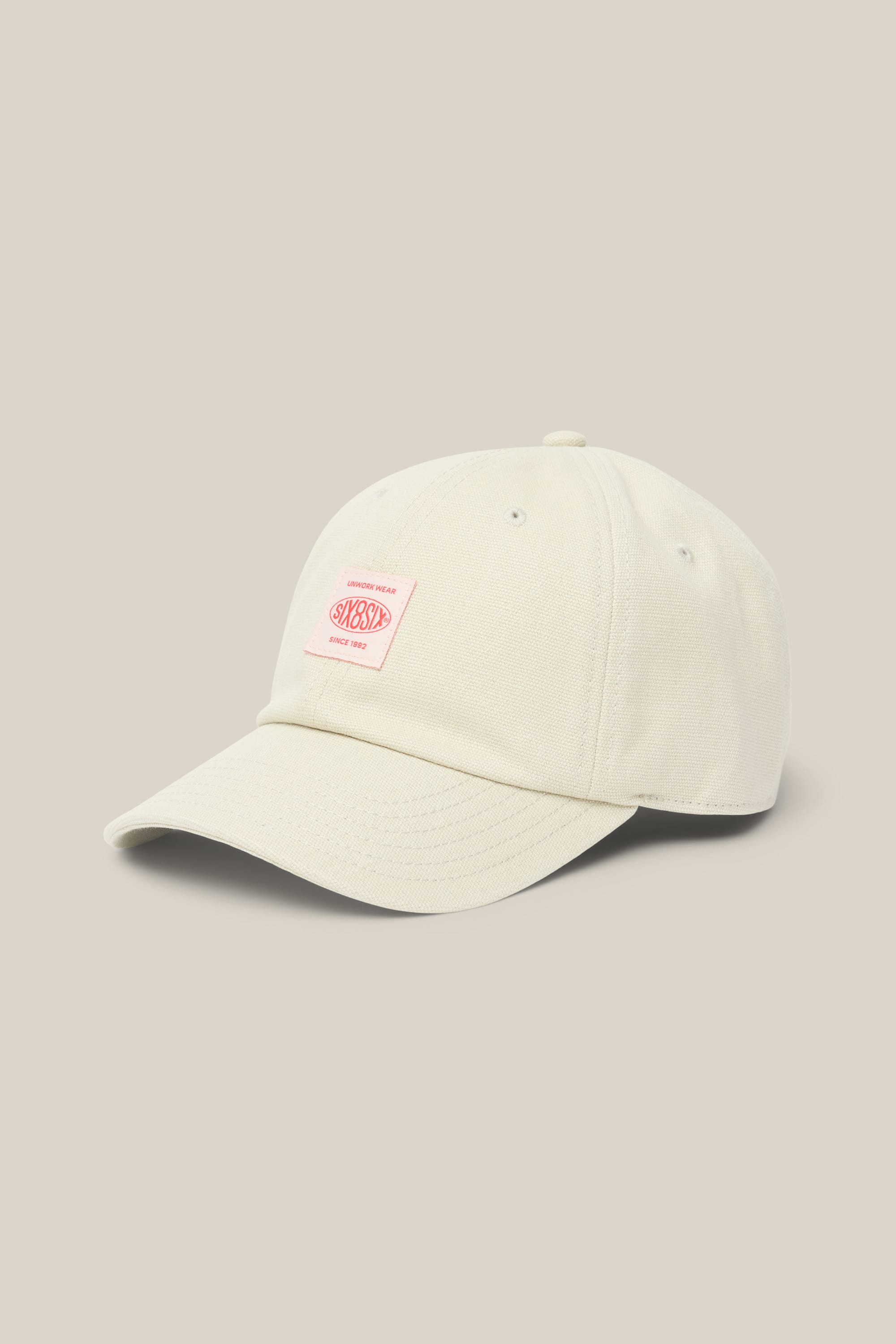 The 686 Low-Pro Unwork Unstructured 6 Panel Hat by 686 is a light beige men's woven hat with a curved brim and a small pink rectangular front patch featuring text, shown on a matching beige background.