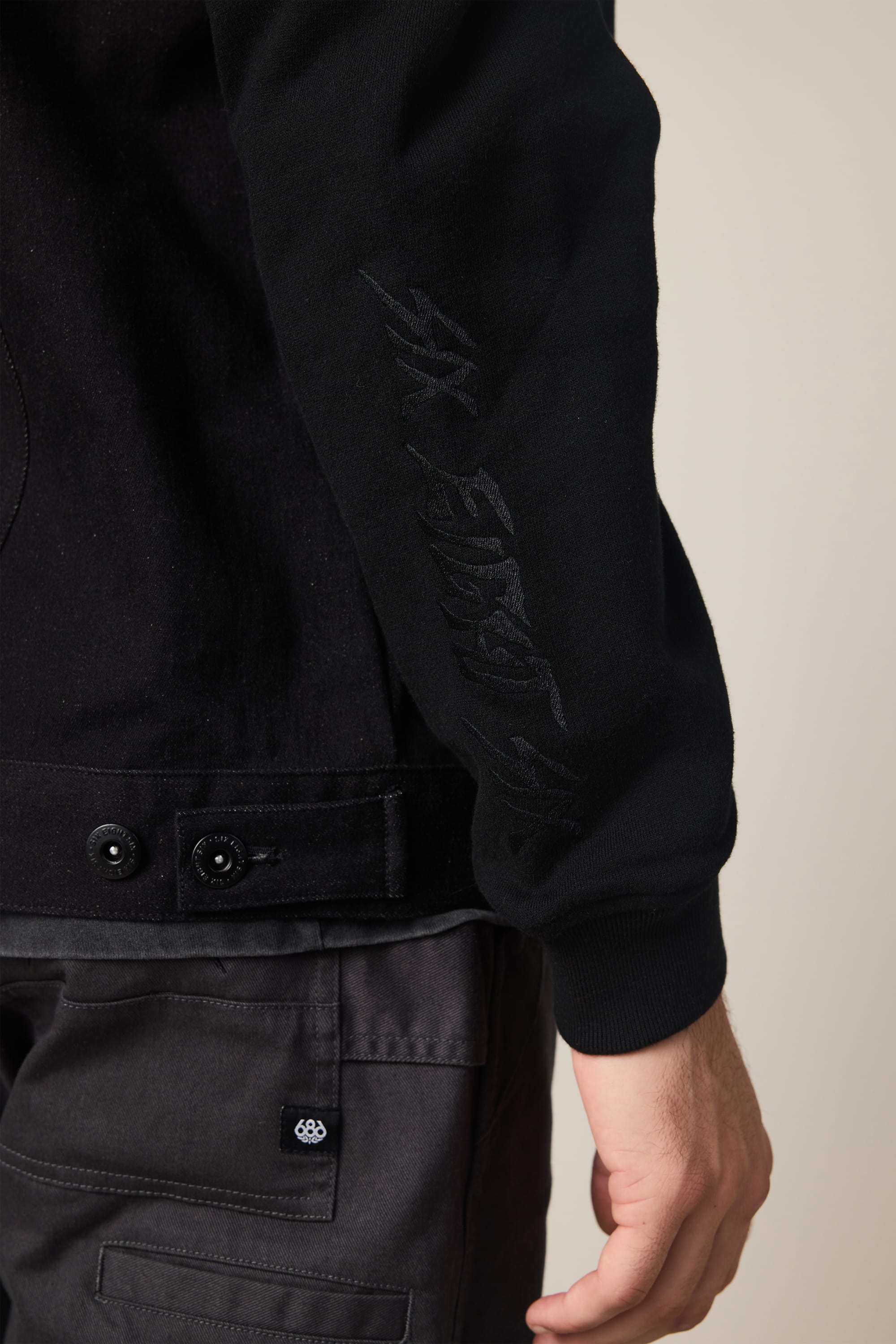 Close-up of a person wearing the 686 Men's Metalhead Trucker Jacket with subtle sleeve embroidery and dark cargo pants. Their hand rests at their side, set against a plain, light background. Brand: 686.