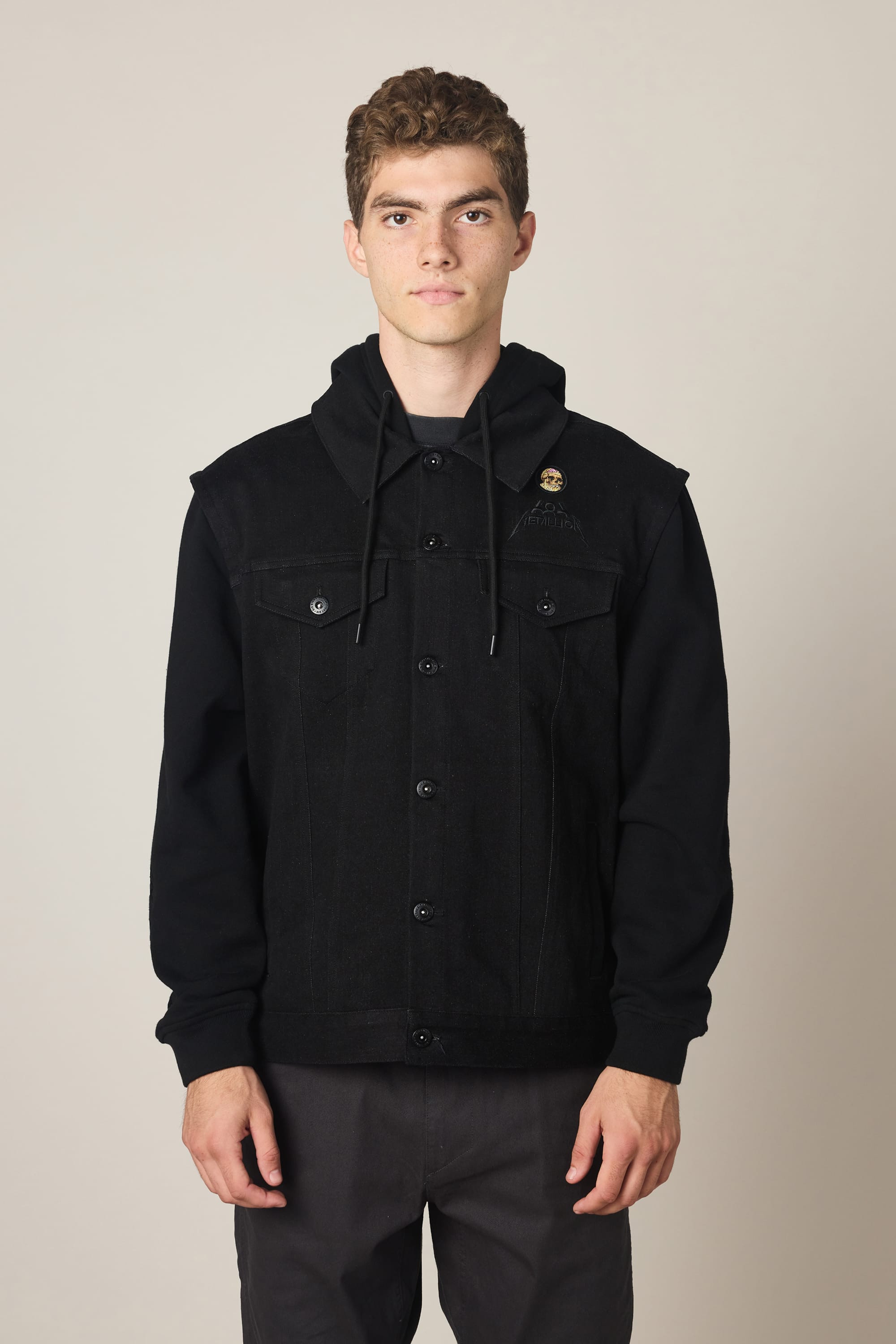 A young man with short curly hair wears the 686 Men's Metalhead Trucker Jacket and dark pants, standing against a plain light background, looking directly at the camera with a neutral expression.