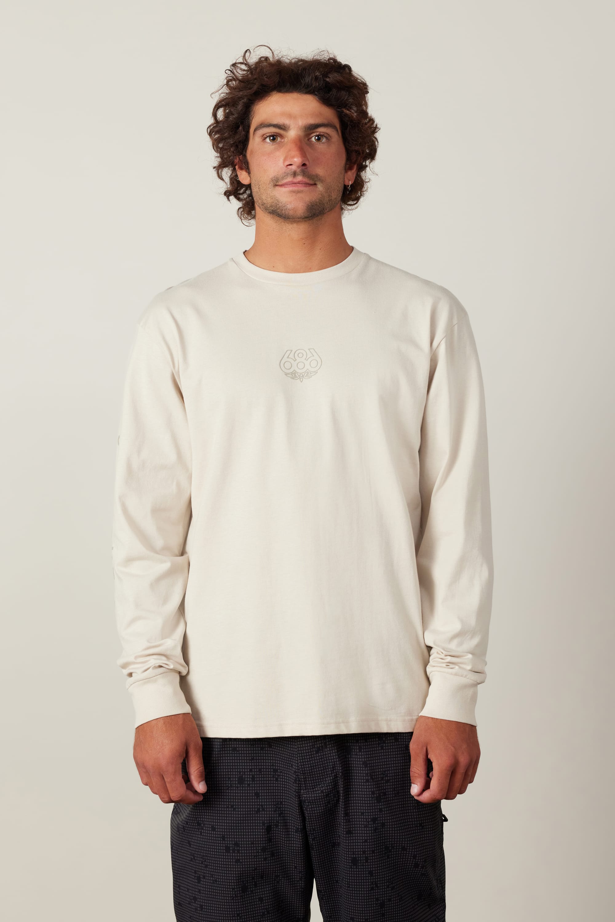 A man with curly brown hair wears the 686 High Def Authentic L/S Tee by 686, an off-white, 100% cotton long-sleeve shirt featuring a small center chest logo, paired with black patterned pants against a plain light background.