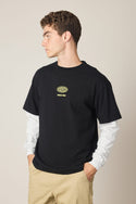 A young man with curly hair looks to the side, wearing the 686 Men's 92 Relaxed LS Double Trouble Tee in black over a white long-sleeve shirt and khaki pants. The tee features a yellow and white circular chest logo.
