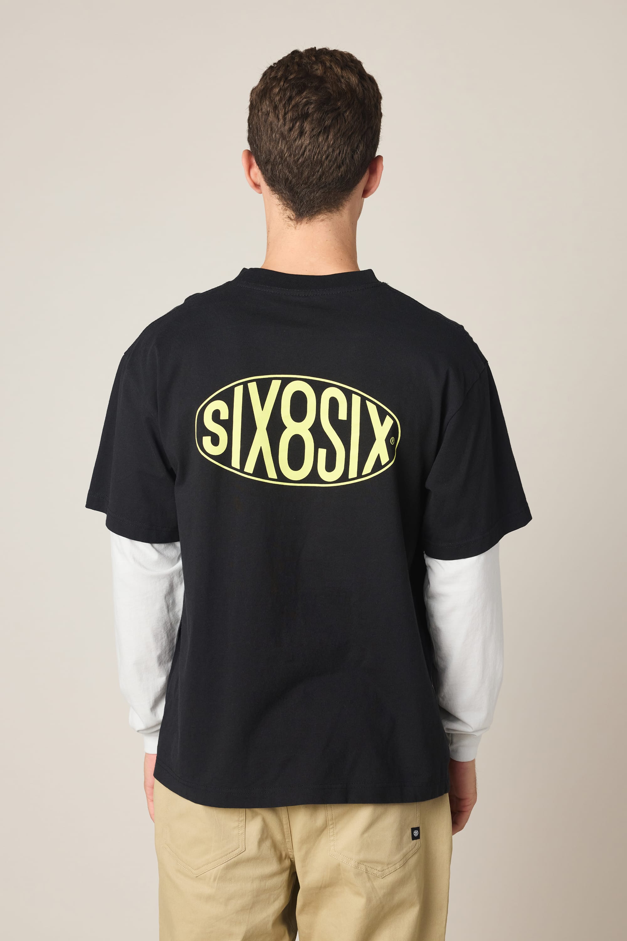 A person faces away, wearing the 686 Men's 92 Relaxed LS Double Trouble Tee in black with "SIX8SIX" in yellow oval on the back, layered over a white long-sleeve shirt and beige pants, set against a plain background.