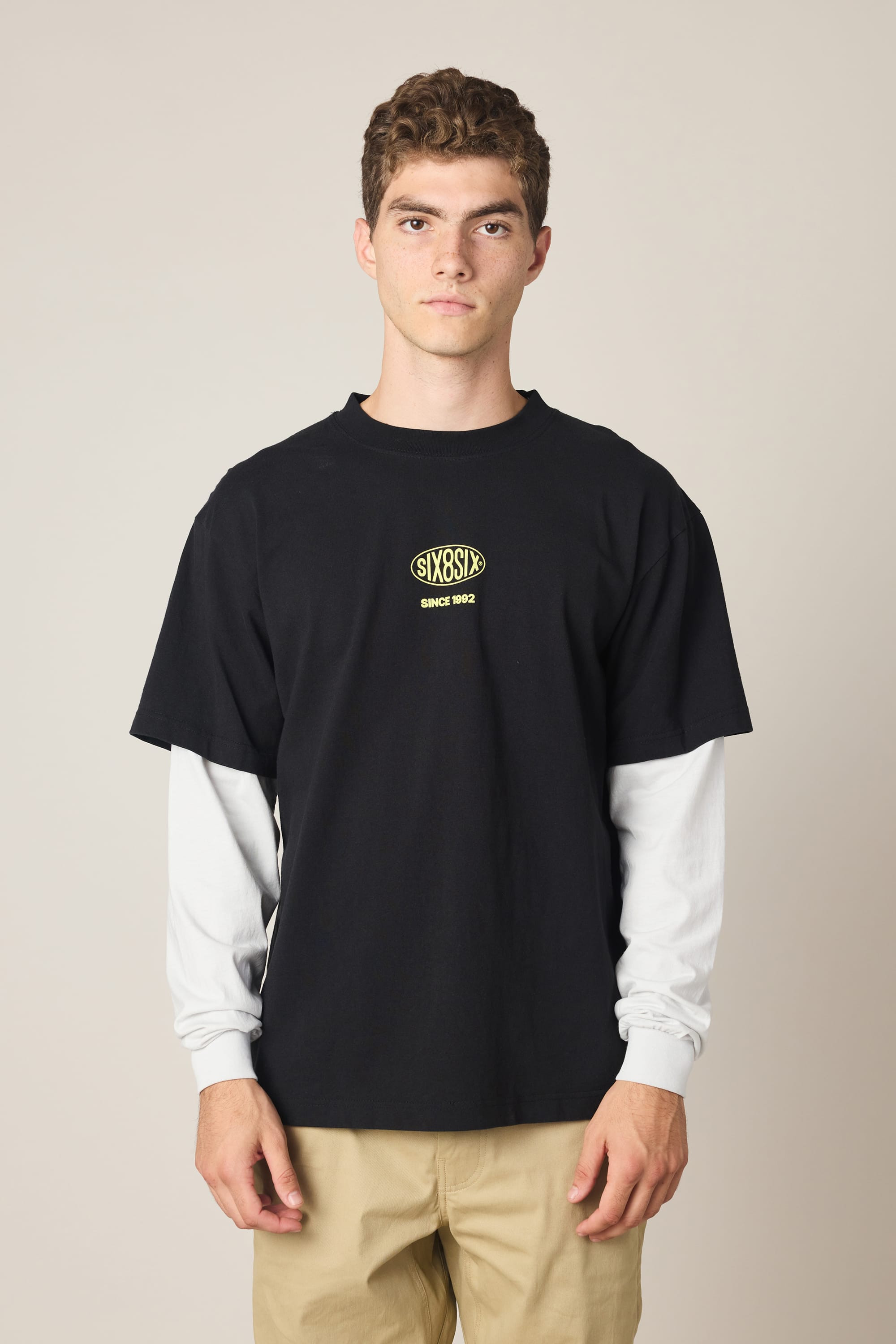 A young man poses against a plain background, wearing the 686 Men's 92 Relaxed LS Double Trouble Tee—100% cotton, black with a yellow graphic and chest text “SIXSIXES SINCE 1992”—layered over a white long-sleeve shirt and beige pants.