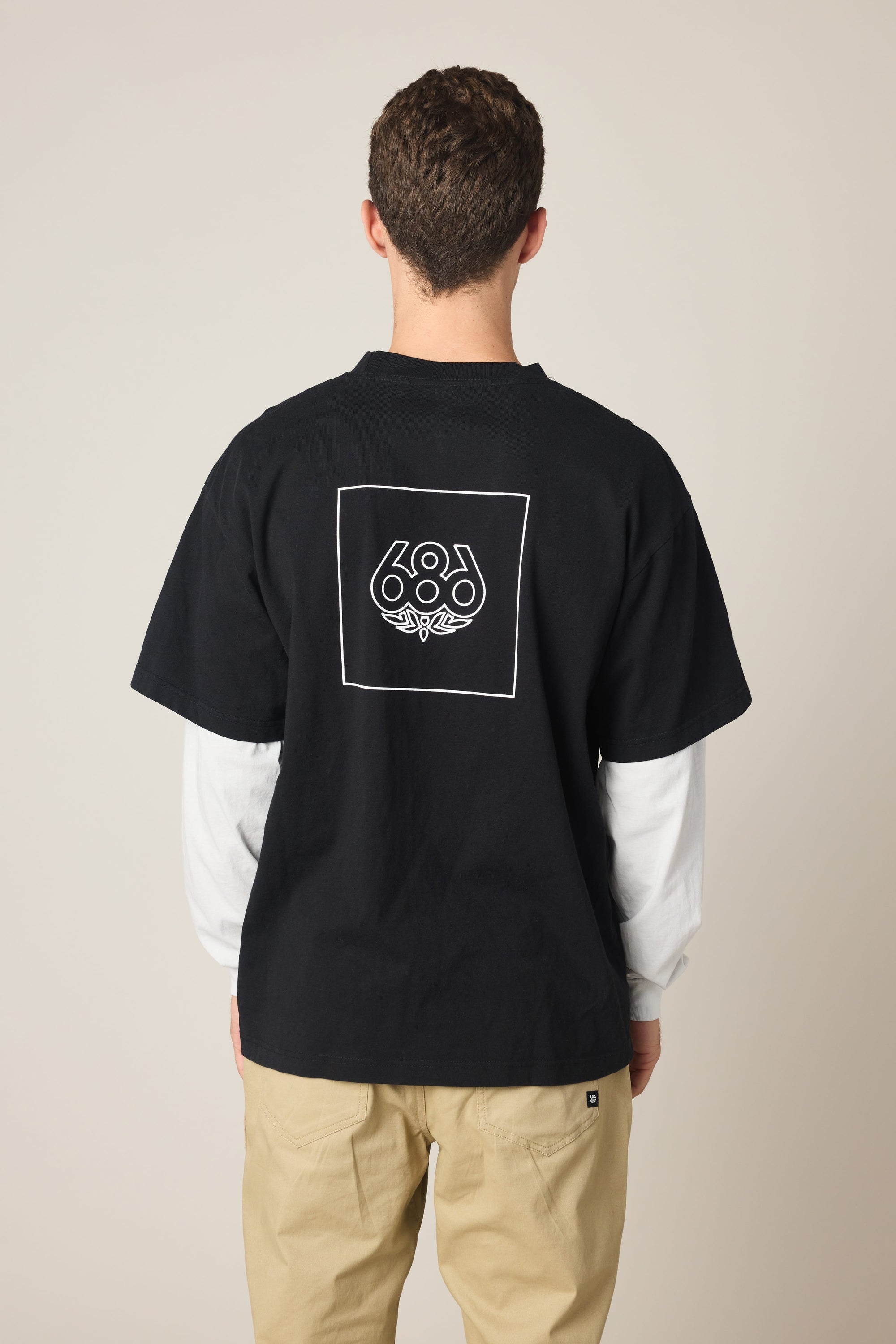 A man faces away, wearing the 686 Wordmark Relaxed 2fer LS Tee—a black cotton t-shirt with a white outlined square and 686 graphic on the back—layered over a white long-sleeve shirt and paired with light beige pants.