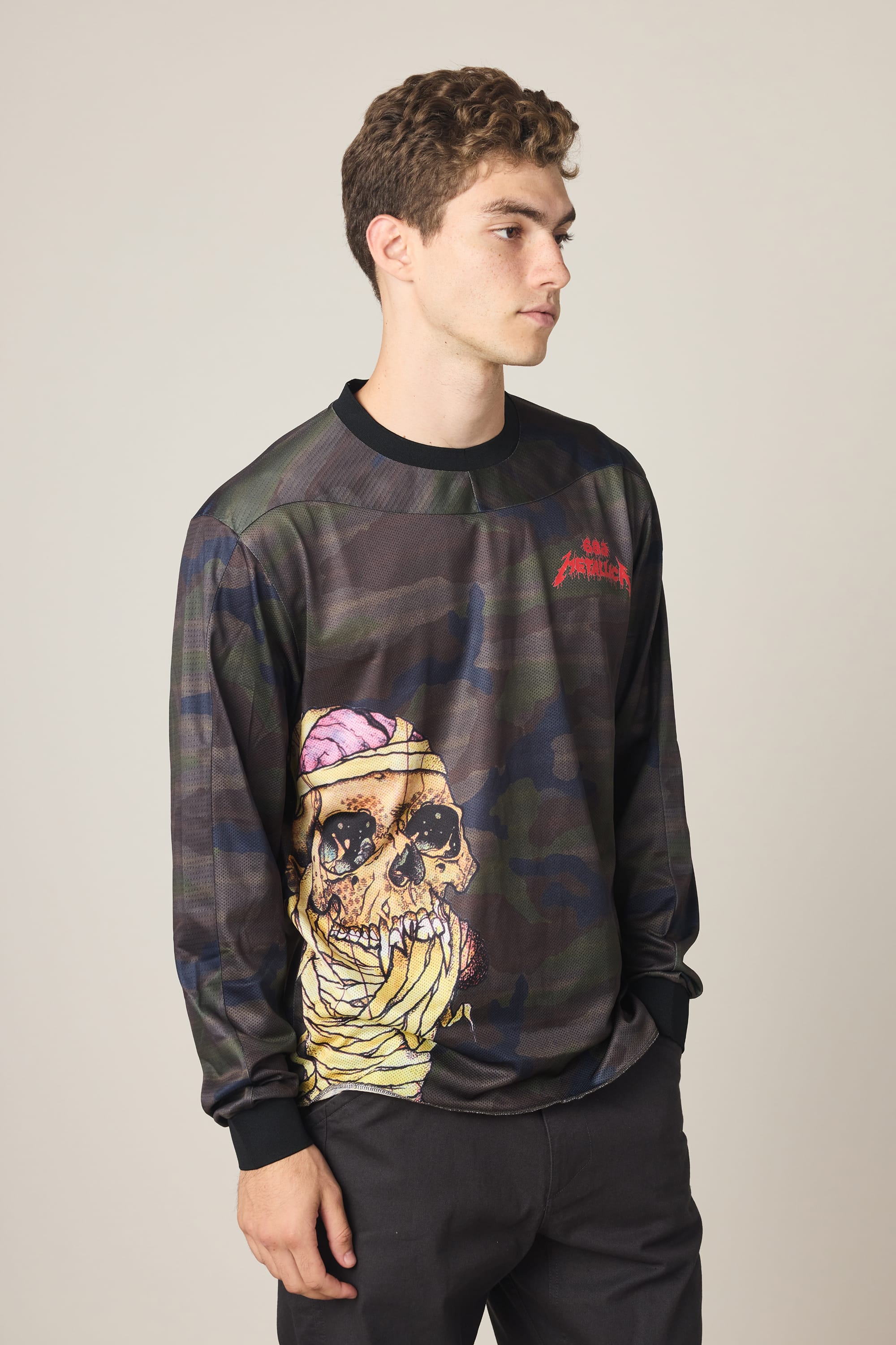 A young man in a 686 Men's Metallica Turbo LS Tech Tee, a dark camo-patterned 100% polyester shirt with a large skull graphic showing a pink brain and yellow accents, stands against a plain light background.