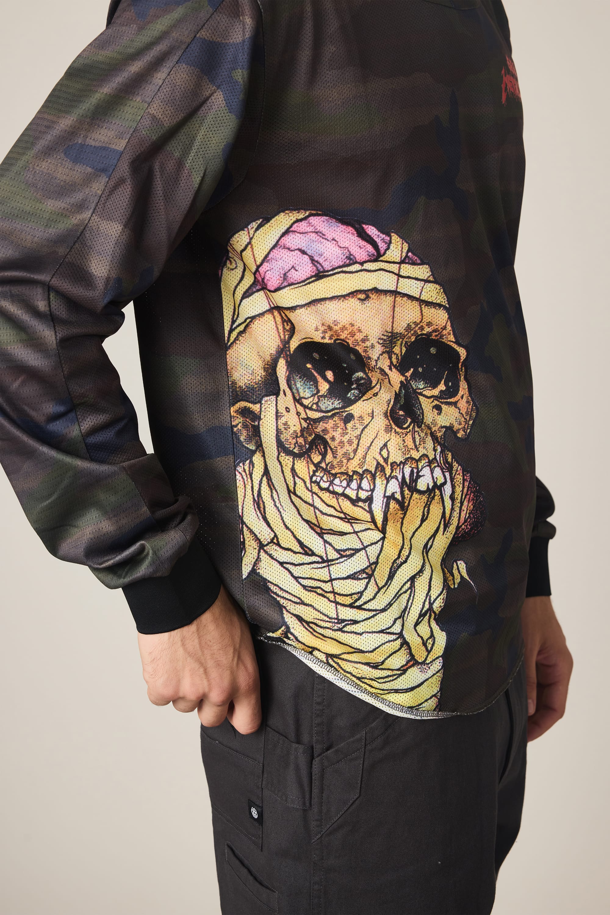 A person wears the 686 Men's Metallica Turbo LS Tech Tee, a men's polyester t-shirt by 686 with camouflage long sleeves and a striking skull graphic, paired with black pants and one hand in their pocket.