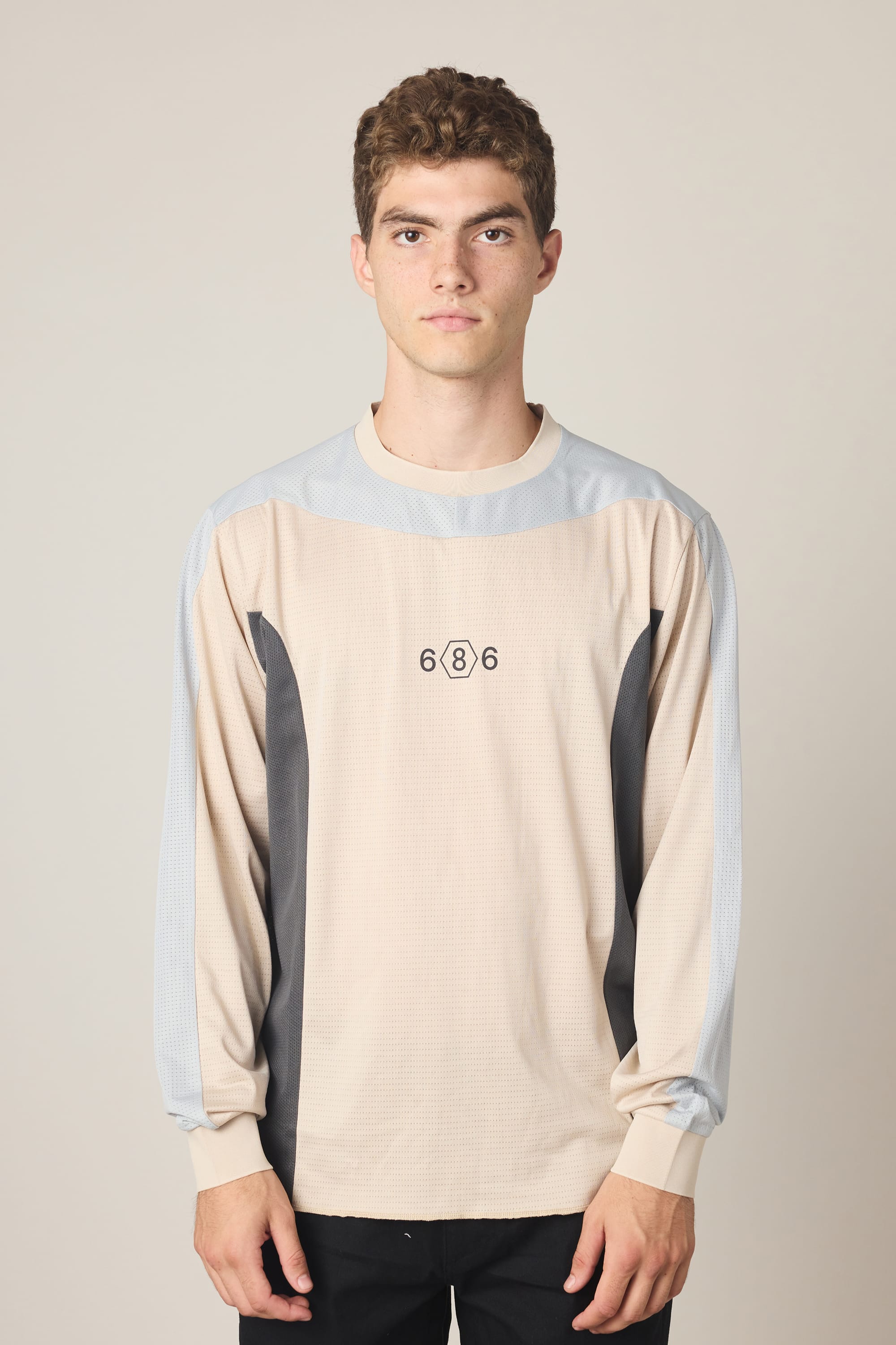 A young man with short curly hair wears the 686 Men's Hex Turbo LS Tech Tee by 686, featuring a beige and gray colorway with "686" on the chest. He stands against a plain light background, looking directly at the camera with a neutral expression.
