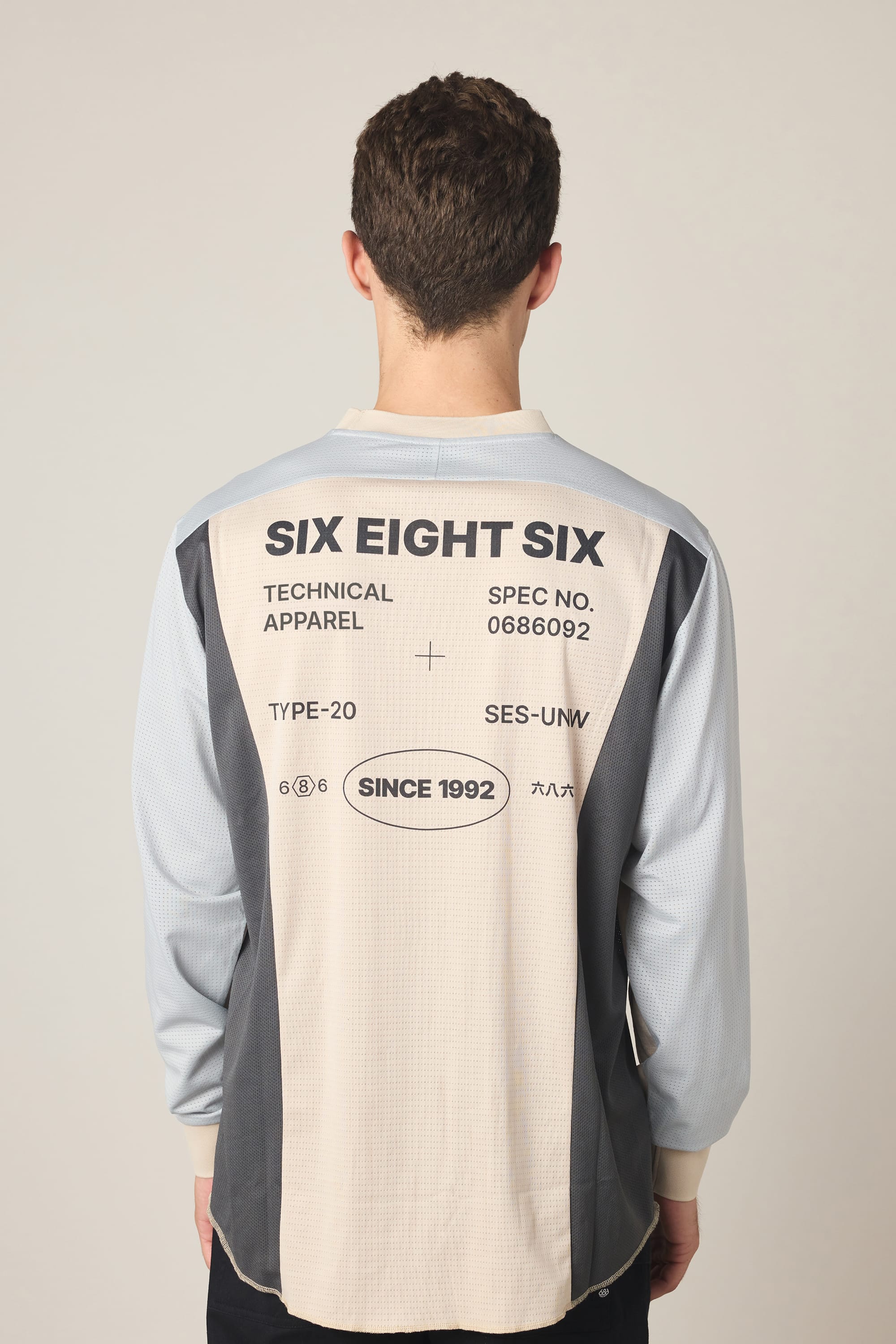 A man with short brown hair, seen from the back, wears the 686 Men's Hex Turbo LS Tech Tee—a long-sleeve color-blocked polyester shirt by 686 featuring text and graphics like SIX EIGHT SIX, TECHNICAL APPAREL, and SINCE 1992.