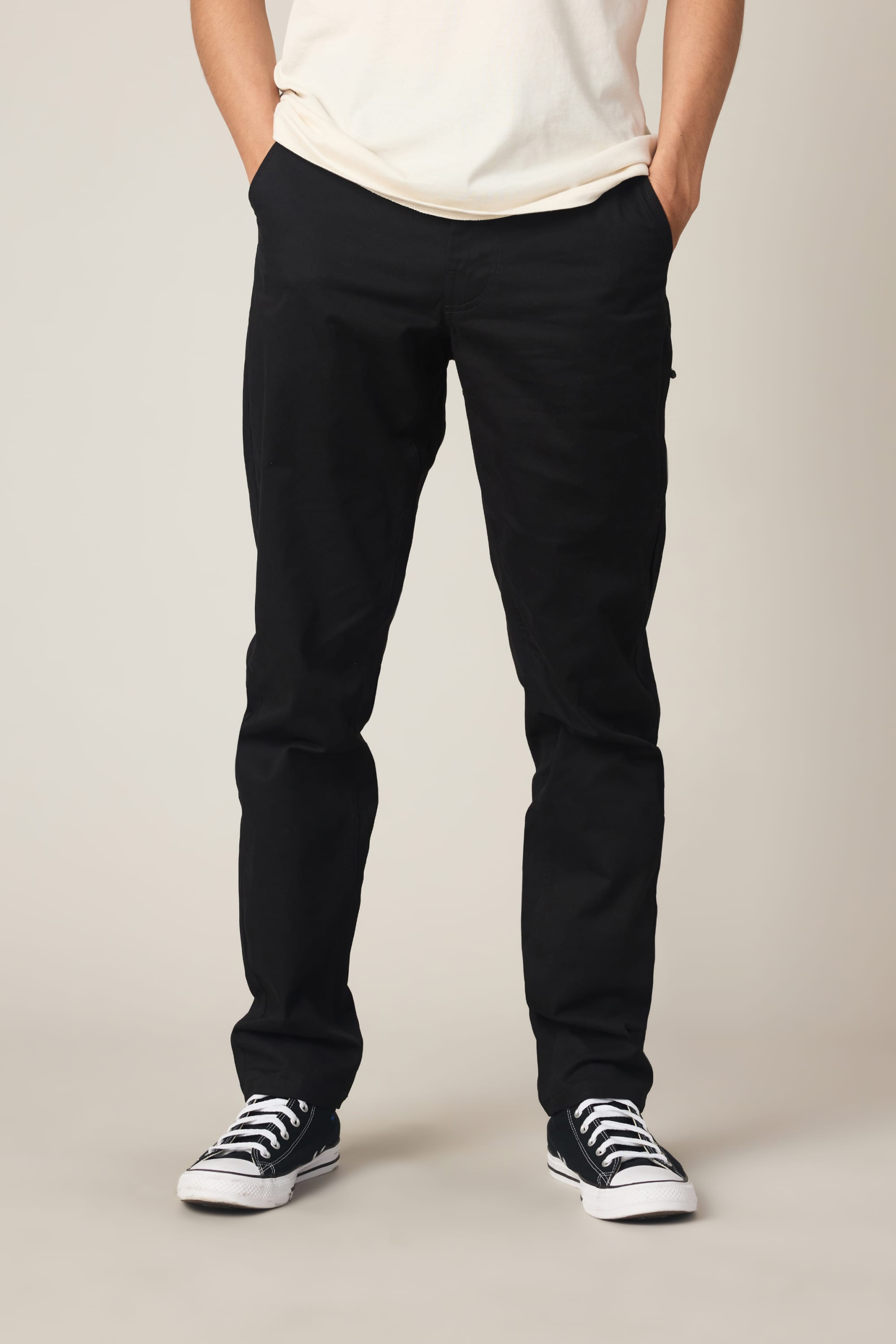 A person stands with hands in their pockets against a plain light background, wearing a white T-shirt, 686 Men's Everywhere® Standard Pant - Slim Fit by 686, and black-and-white sneakers. Their head is not visible in the image.