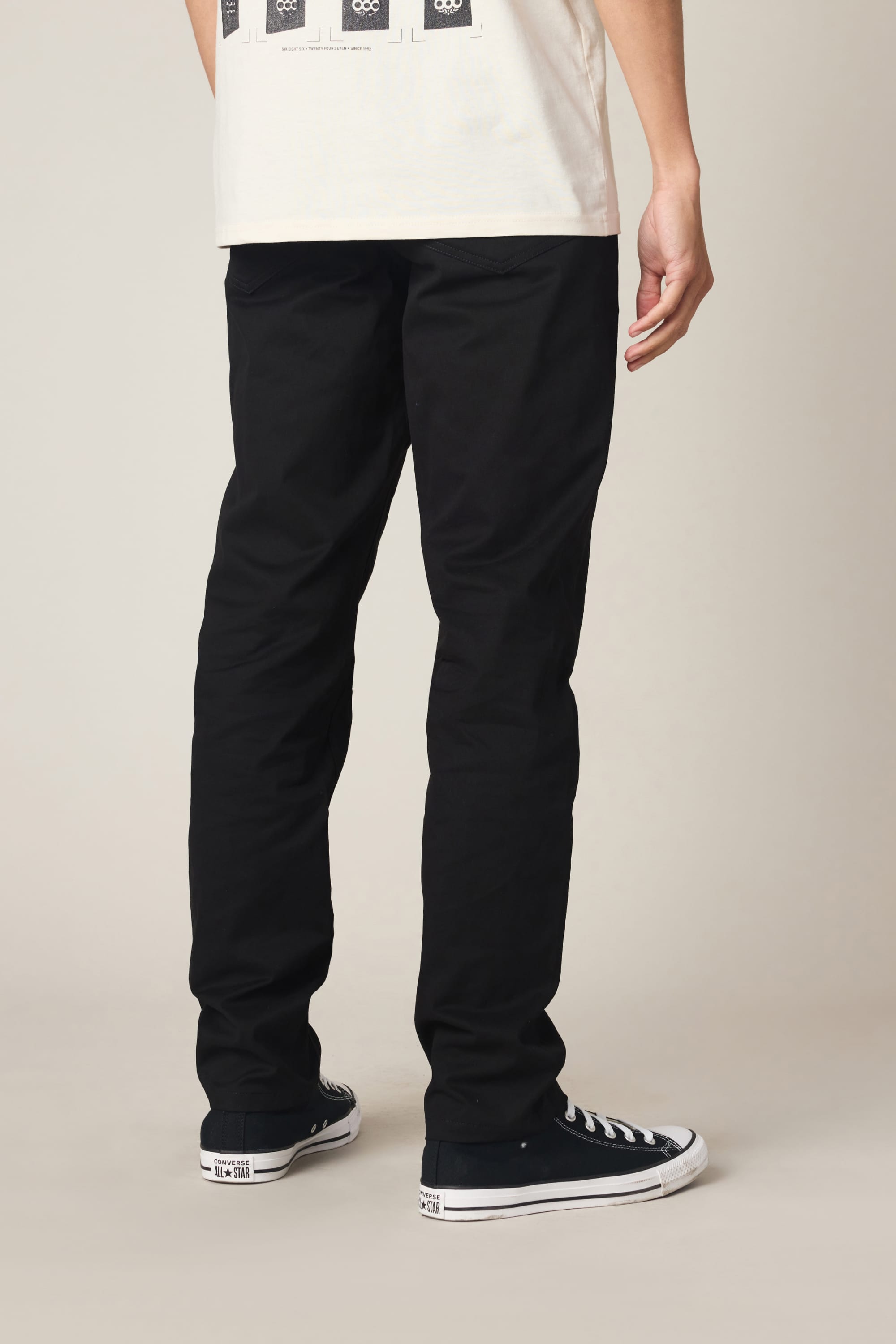 A person stands facing away, only their lower half visible against a plain background, wearing black 686 Men's Everywhere® Standard Pant - Slim Fit along with a white t-shirt and black low-top sneakers featuring white soles.