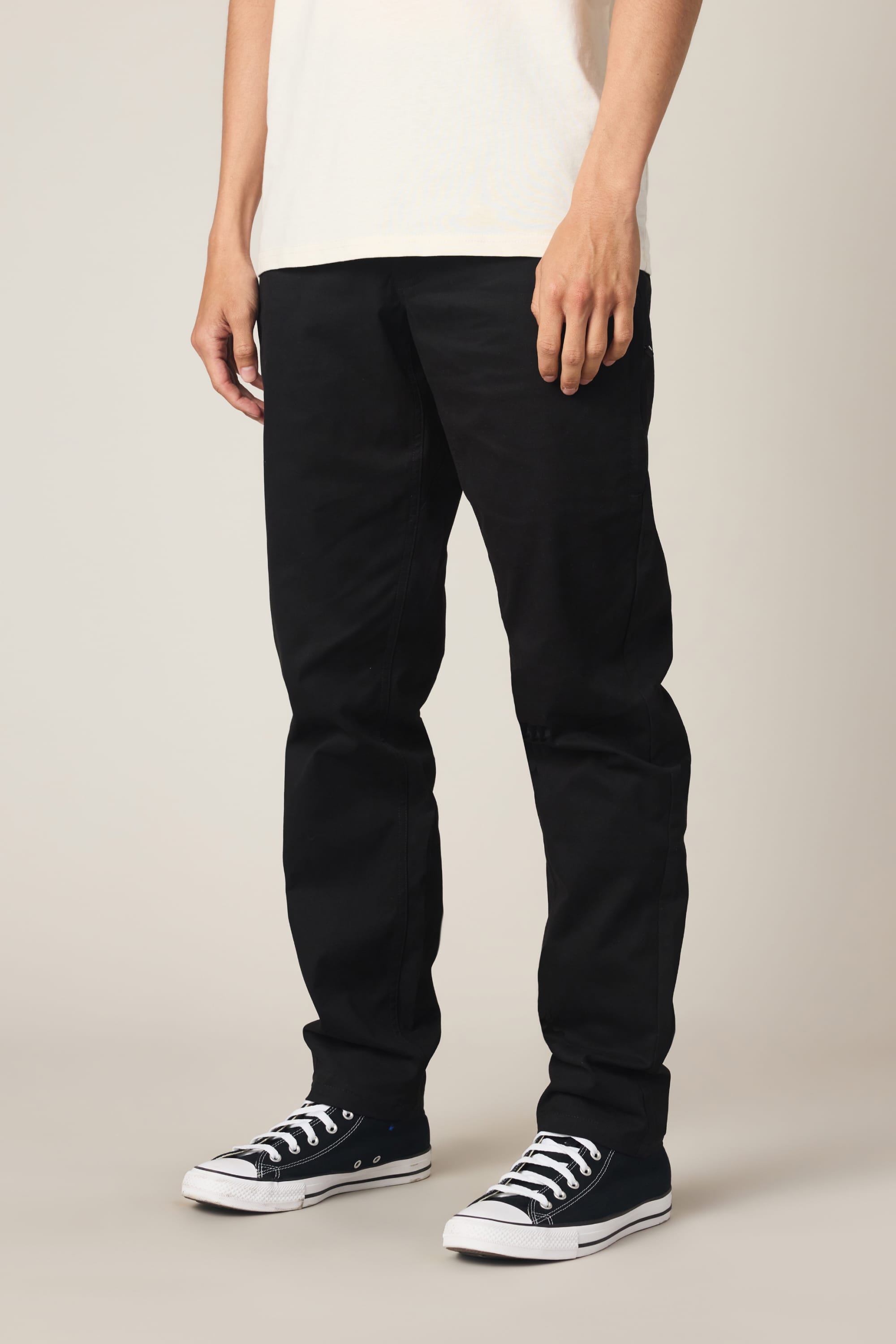 A person wearing 686 Men's Everywhere® Standard Pant - Slim Fit in black, paired with a white shirt and black low-top sneakers with white soles, stands against a plain light background. Only the lower half of the body is visible.
