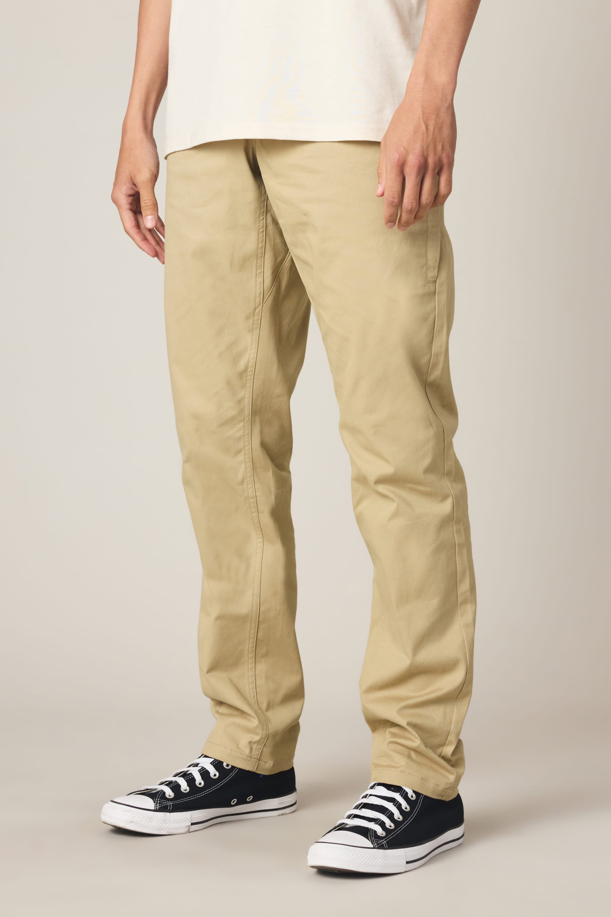 A person wearing 686 Men's Everywhere® Standard Pant - Slim Fit in tan khaki, a white shirt, and black low-top Converse sneakers stands against a plain light background. Only the lower half of the body is visible.
