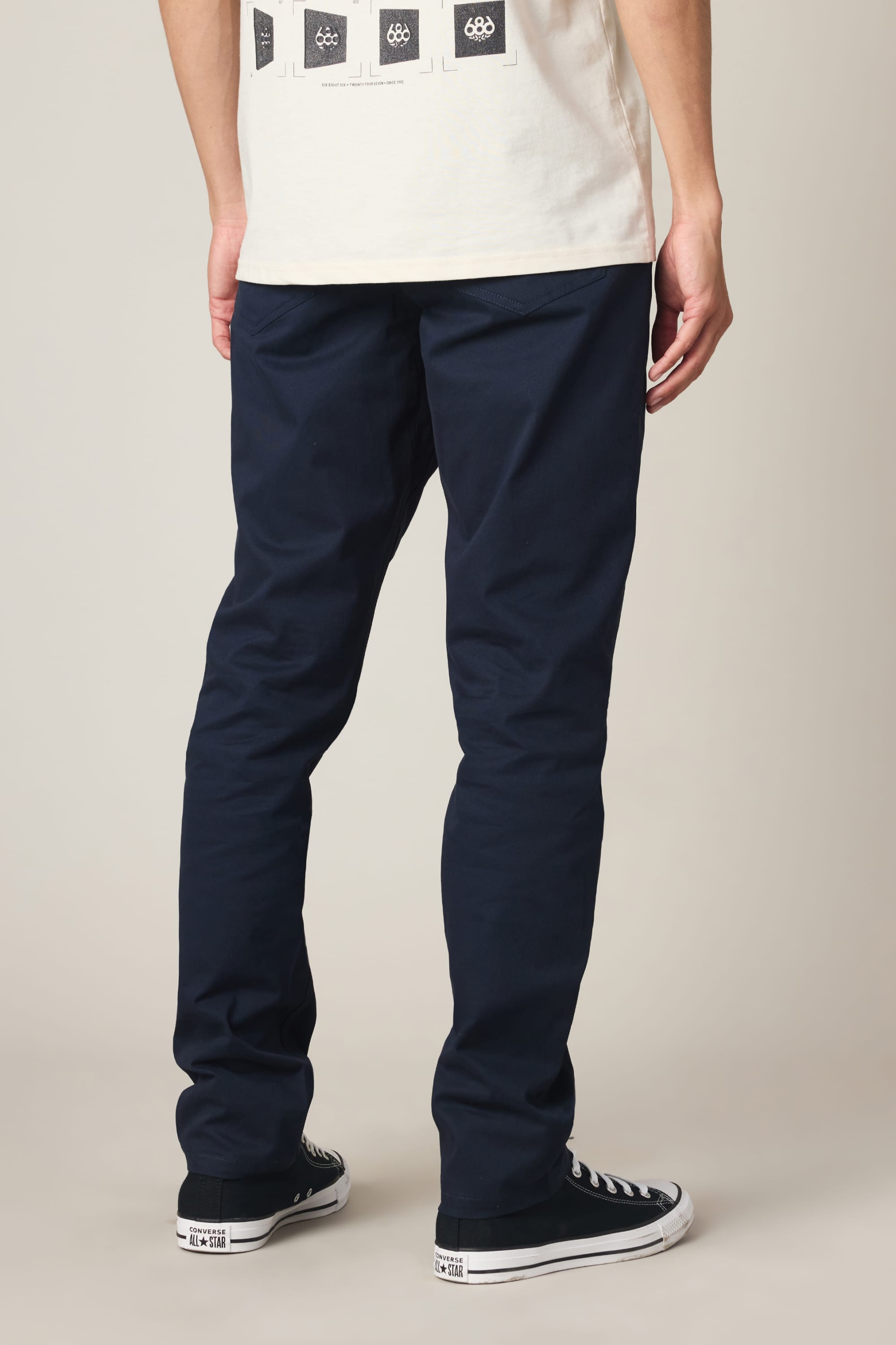 A person stands facing away, wearing a white T-shirt with back graphics, 686 Men's Everywhere® Standard Pant - Slim Fit in navy blue by 686, and black low-top sneakers with white soles against a plain background.