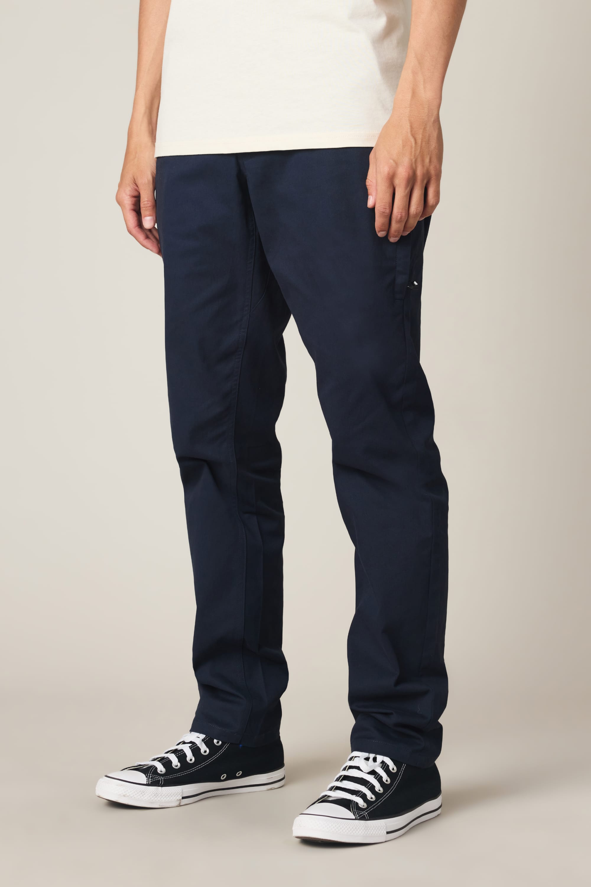 A person stands against a plain light background, shown from shoulders down, wearing a white t-shirt, 686 Men's Everywhere® Standard Pant - Slim Fit in navy blue, and black low-top Converse sneakers.