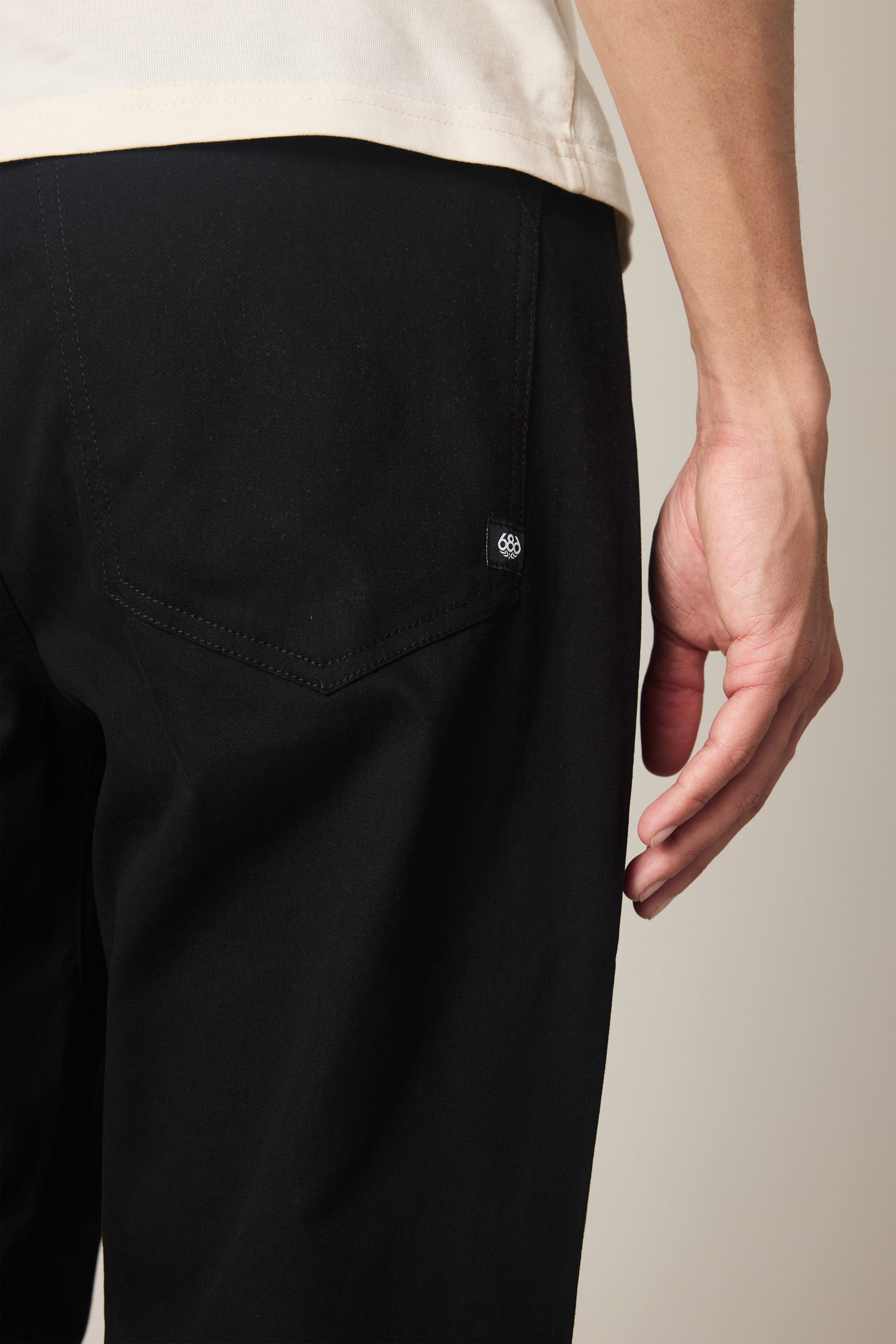 A close-up of a person’s lower back and hand wearing 686 Men's Everywhere® Standard Pant - Relaxed Fit in black, showing the visible back pocket with a small black and white logo tag, paired with a light-colored shirt.