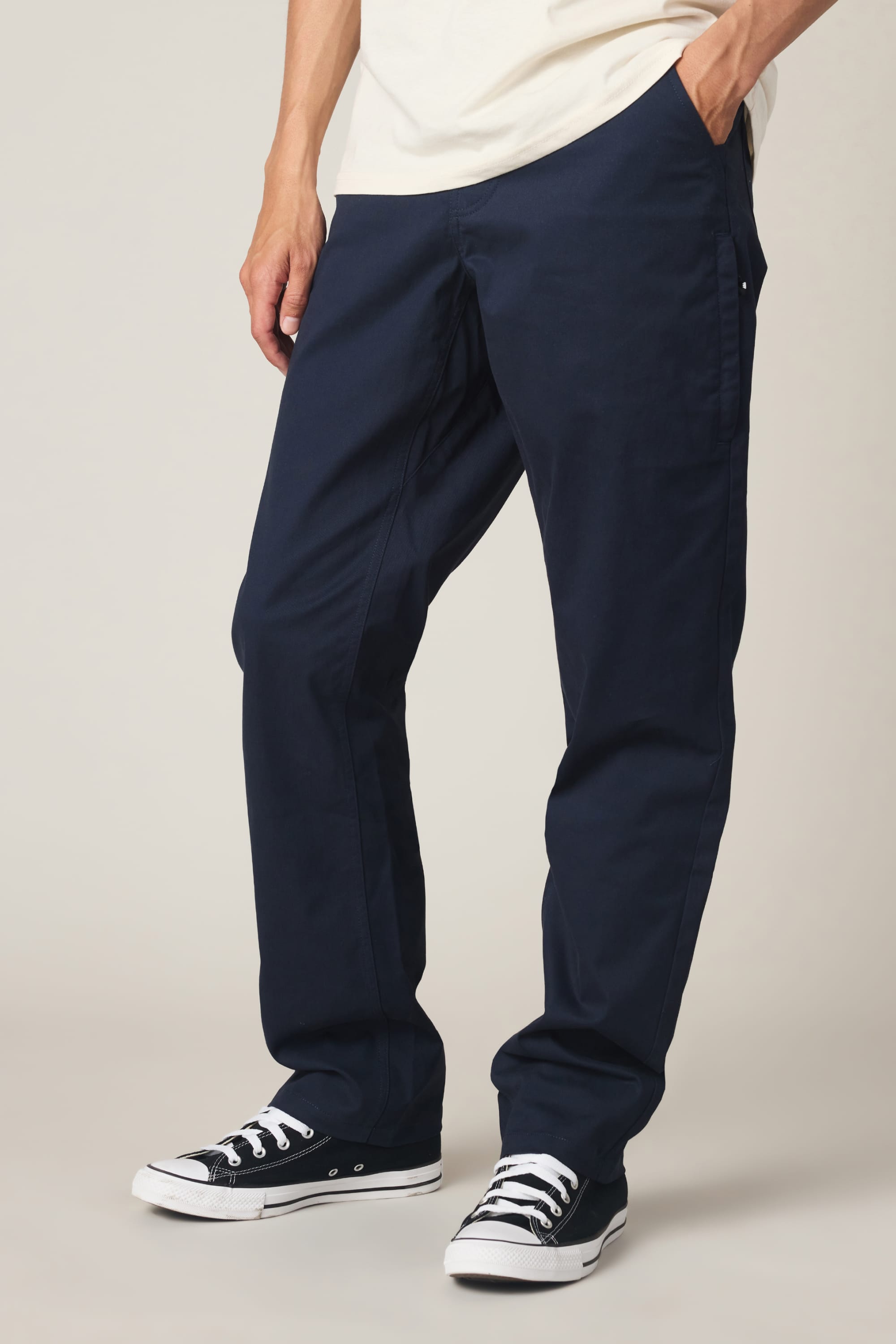 A person wearing 686 Men's Everywhere® Standard Pant - Relaxed Fit in dark blue, a cream t-shirt, and black low-top sneakers stands with one hand in their pocket against a neutral background.
