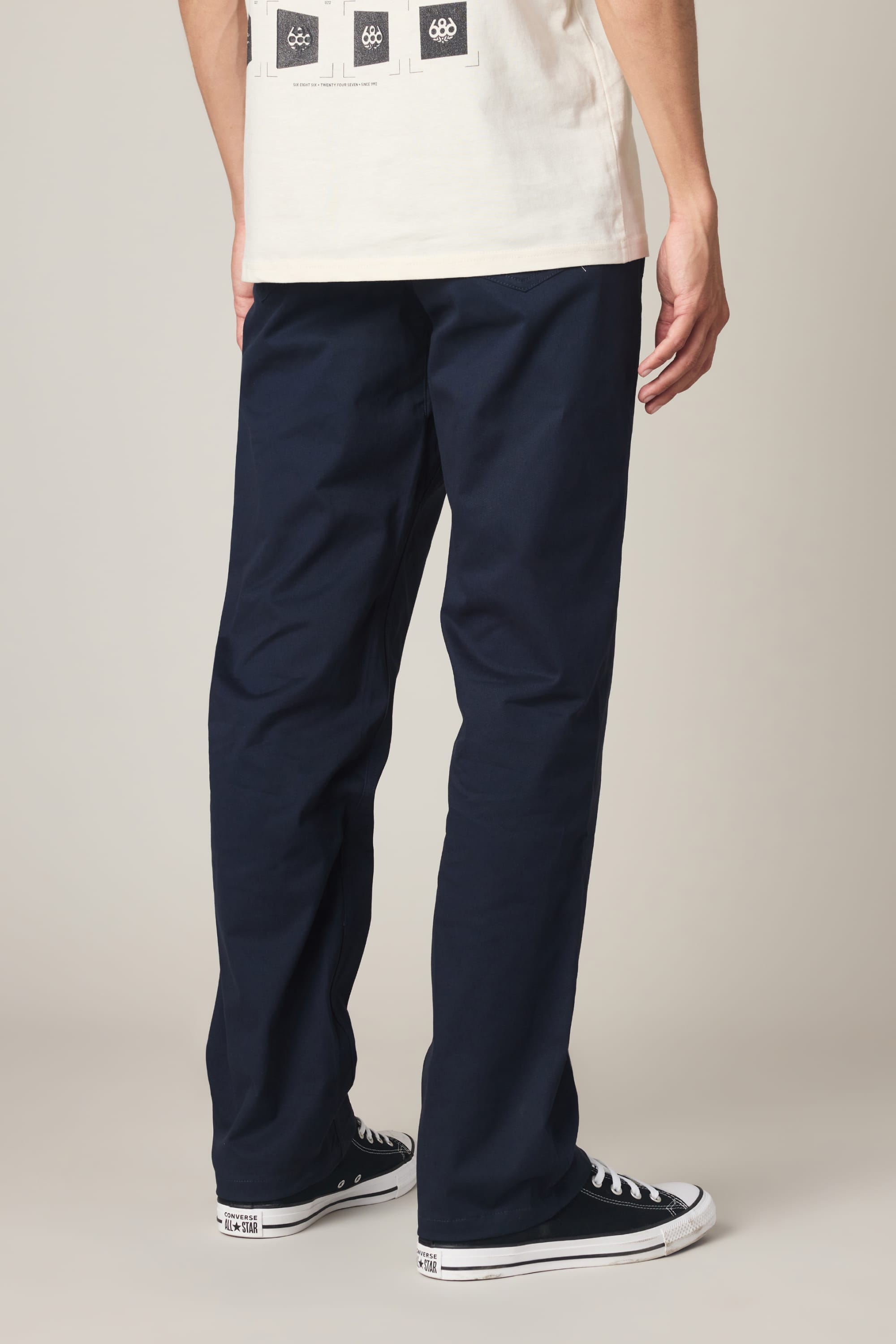A person faces away, wearing 686 Men's Everywhere® Standard Pant - Relaxed Fit in dark blue by 686, a white graphic-back tee, and black-and-white sneakers against a plain background.