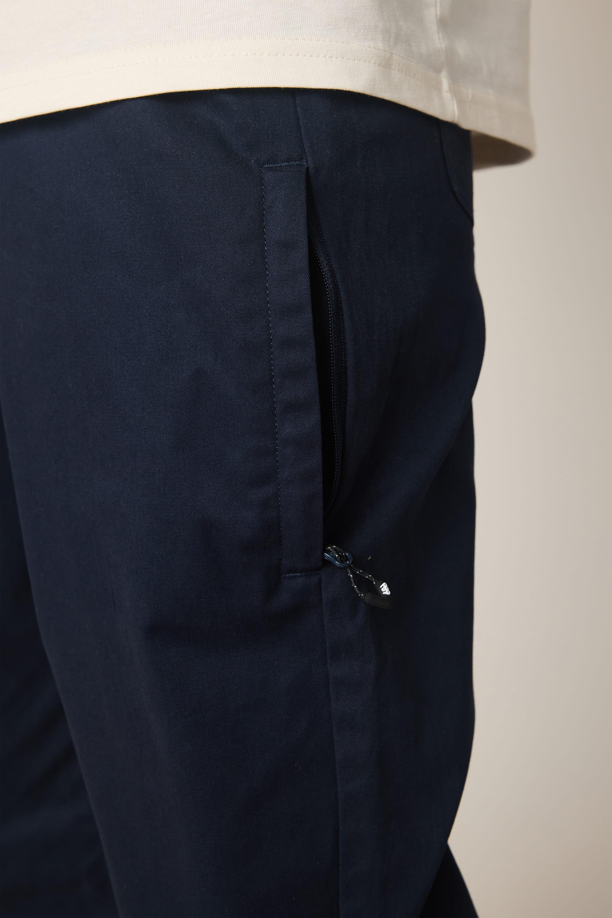 Close-up of a person wearing the 686 Men's Everywhere® Standard Pant - Relaxed Fit in dark blue nylon with a zippered side pocket, paired with a light beige shirt.