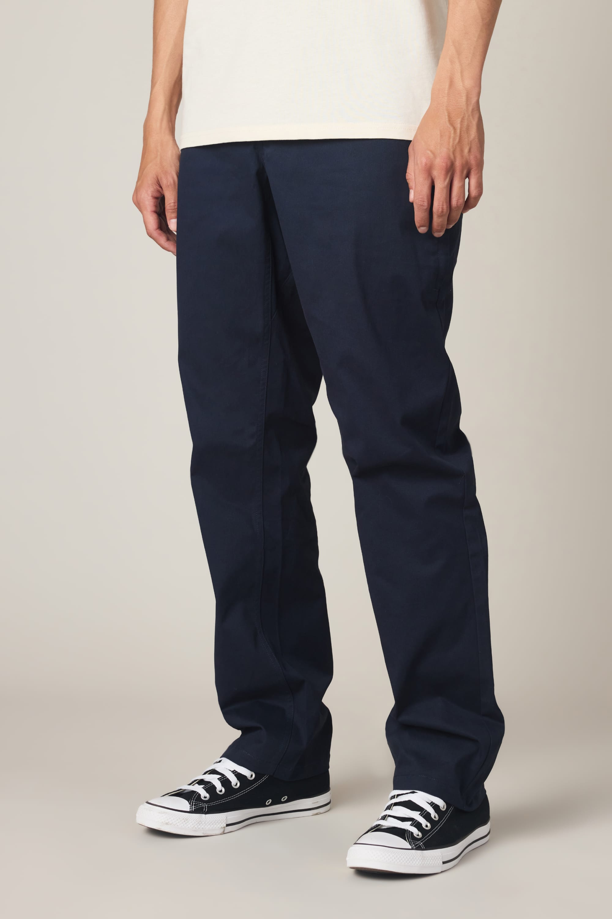 A person wearing 686 Men's Everywhere® Standard Pant - Relaxed Fit in dark blue, a white shirt, and black low-top sneakers with white laces stands against a plain light background.