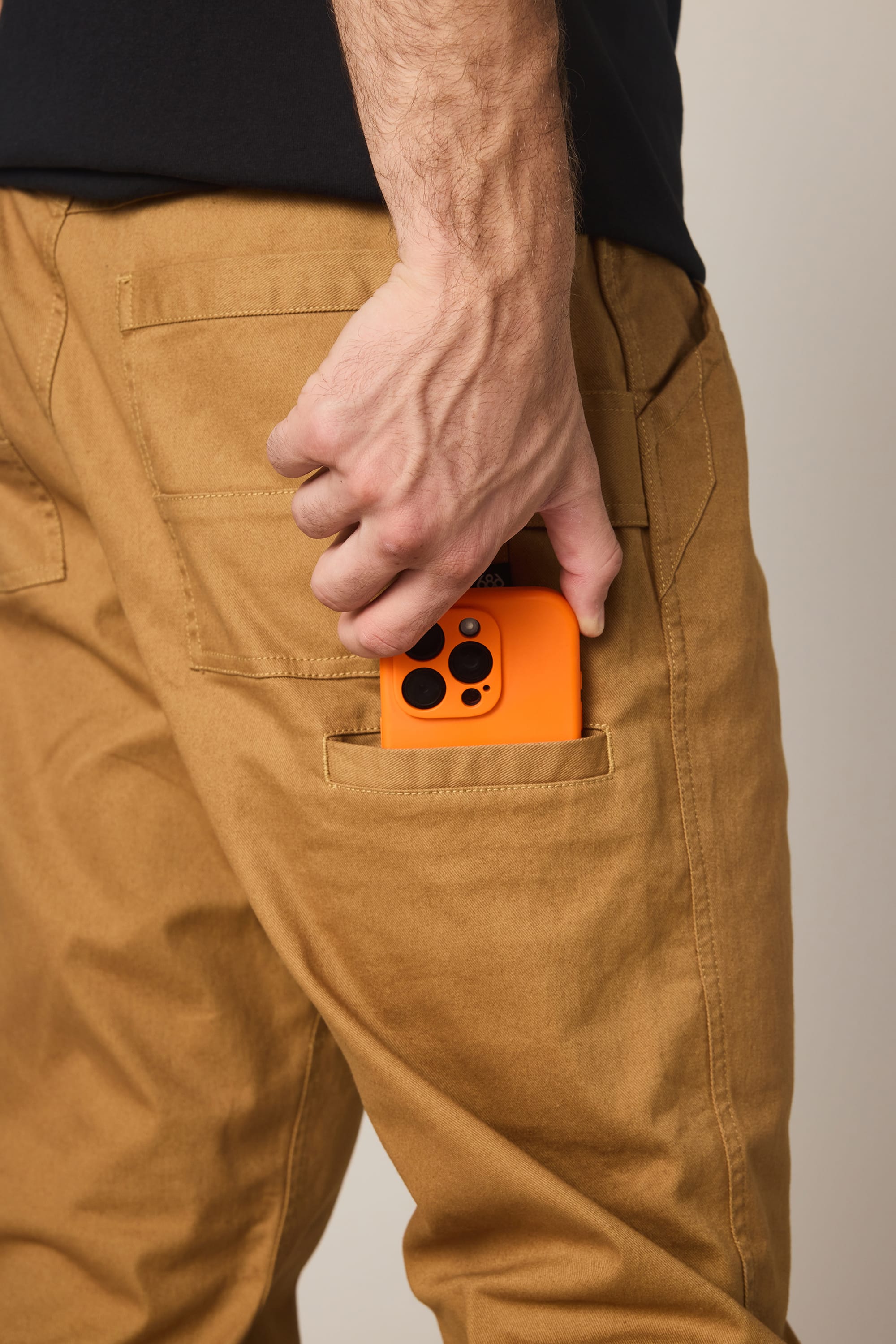 A person wearing 686 Men's Unwork® Tradesman Pant - Relaxed Fit slips an orange smartphone with a black case into the back pocket, their hand partially covering the phone.